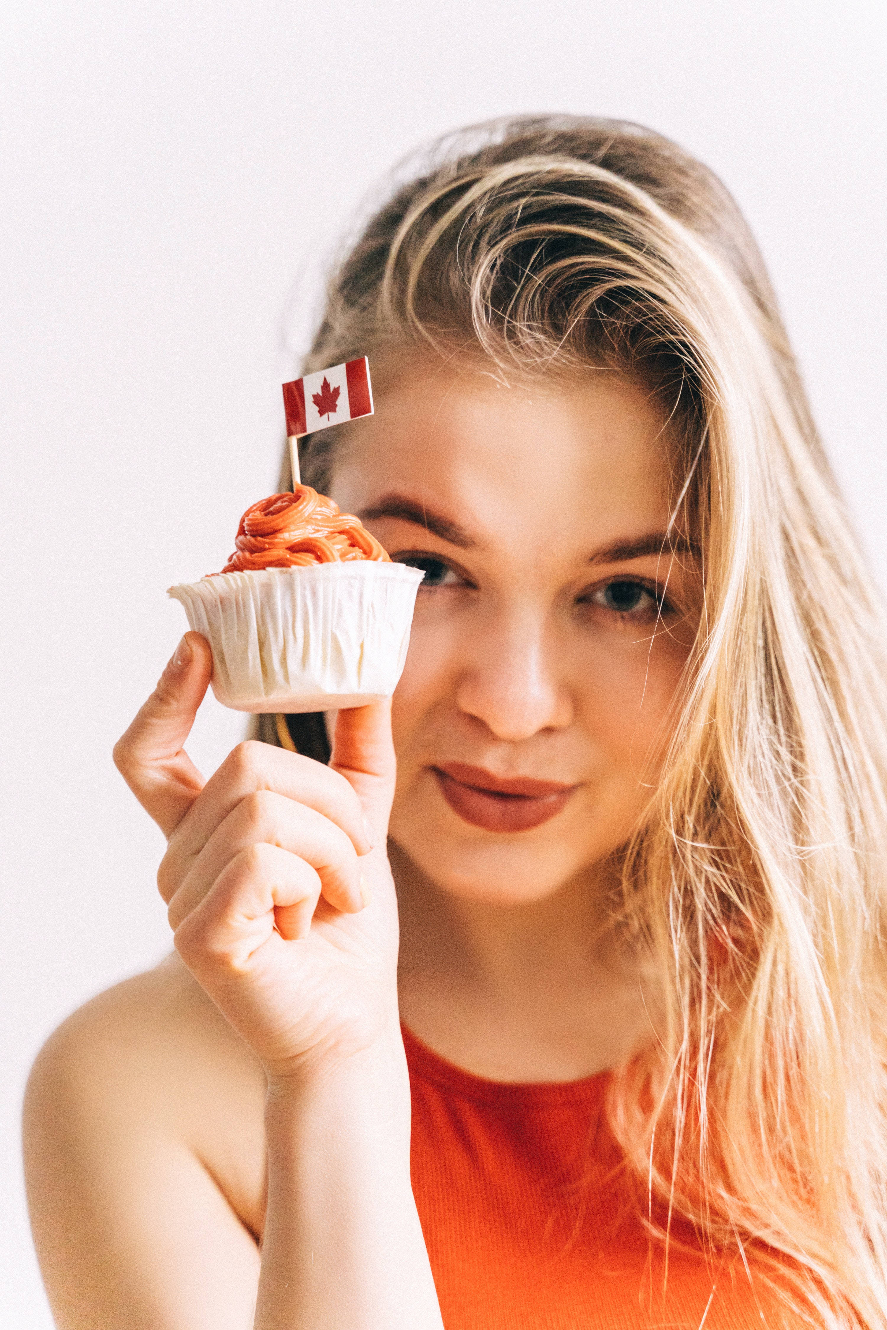 food iPhone 桌布 壁紙 — 一位金髮年輕女性手持一個頂部有細小加拿大國旗的杯子蛋糕，背景為純白色。