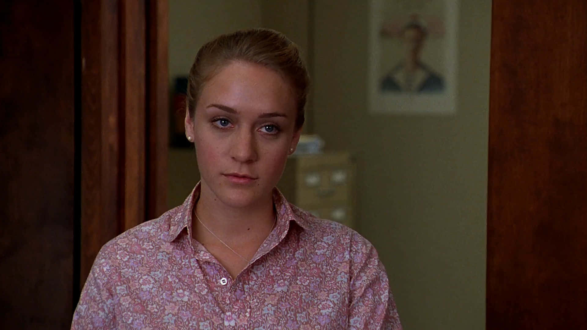 Chloë Sevigny 桌布 壁紙 — 一位金髮年輕女性穿著粉紅色花卉襯衫，站在光線溫暖的室內走廊，直視鏡頭，表情嚴肅。