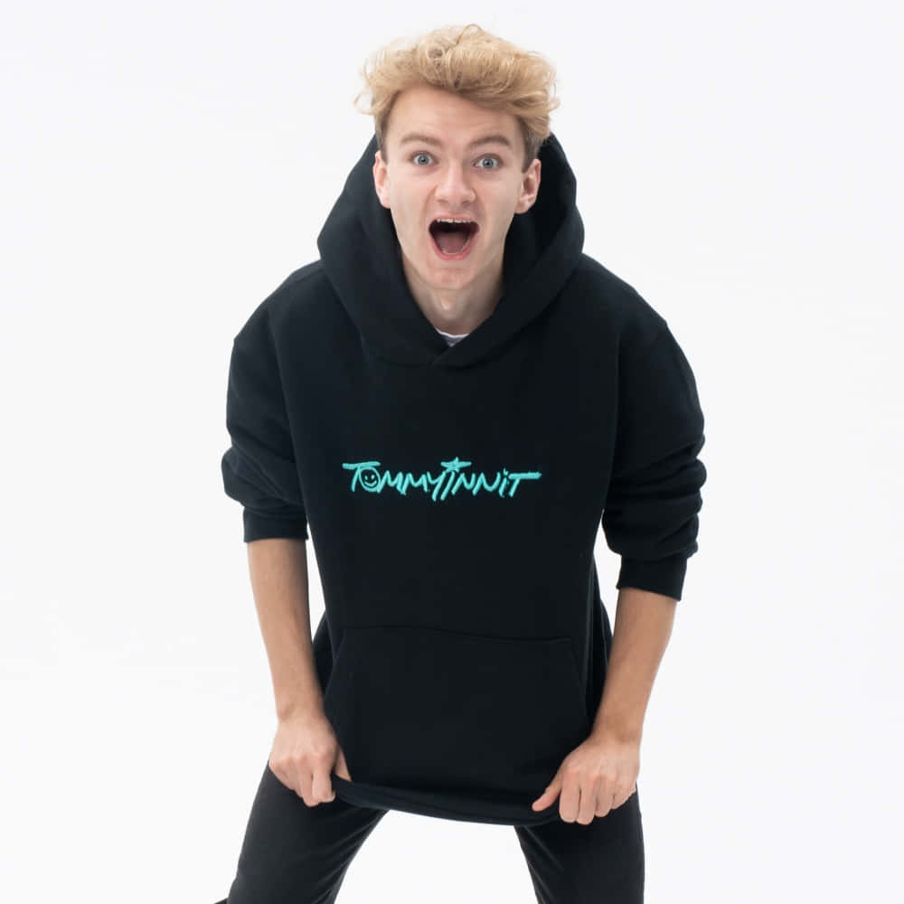 Tommyinnit 桌布 壁紙 — 一位金髮年輕男子穿著帶有發光青綠色標誌的黑色連帽衫，在白色背景前做出誇張的驚訝表情。