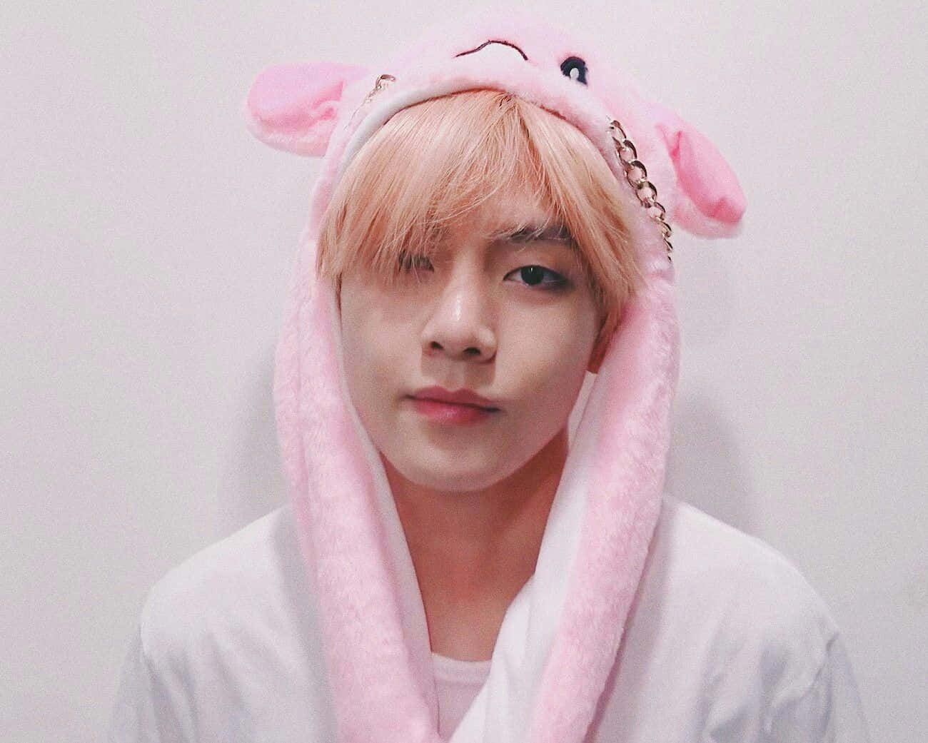 Taehyung Cute 桌布 壁紙 — 一位金髮年輕男子穿著毛茸茸的粉紅色豬主題連帽衫，帶有耳朵和鏈條細節，背景是純白牆壁。