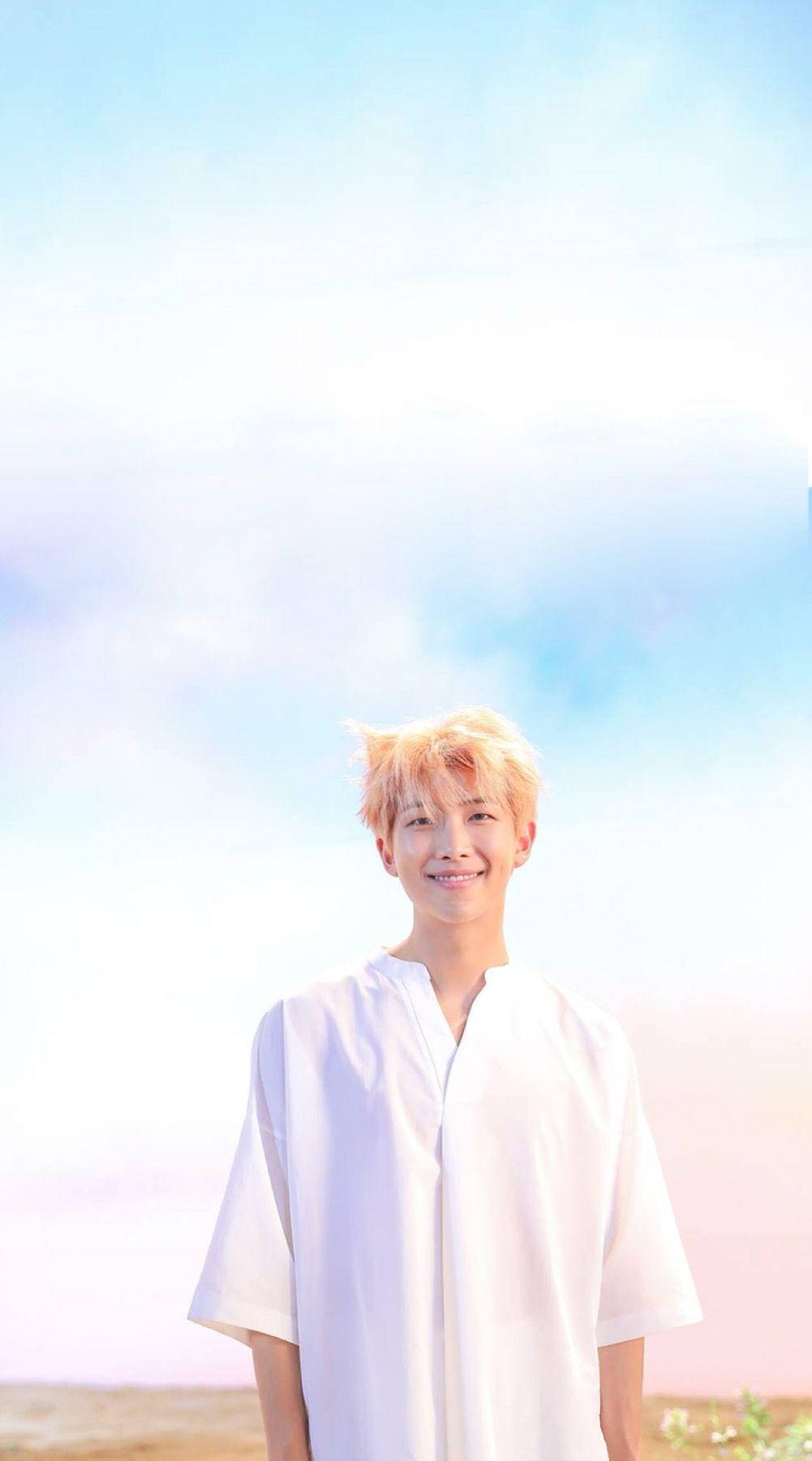RM BTS《Memories 2017》 壁紙 — 一位金髮年輕男子穿著白襯衫，在柔和粉彩天空與輕柔雲朵背景下溫暖微笑。