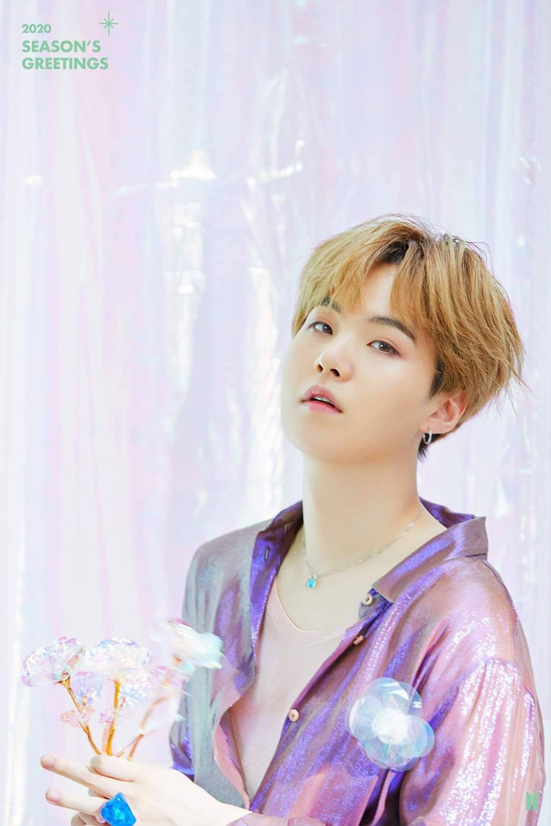 BTS Suga 2020 壁紙 — 一位金髮年輕男子穿著閃亮紫色襯衫，在柔和的粉紅色和白色背景前握著發光的花朵。