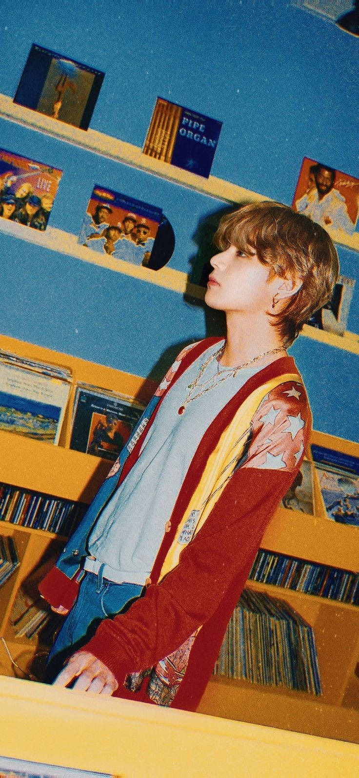 bts photoshoot 桌布 壁紙 — 一位金髮年輕男子靠在藍色牆壁唱片店內裝滿黑膠唱片的黃色架子上。
