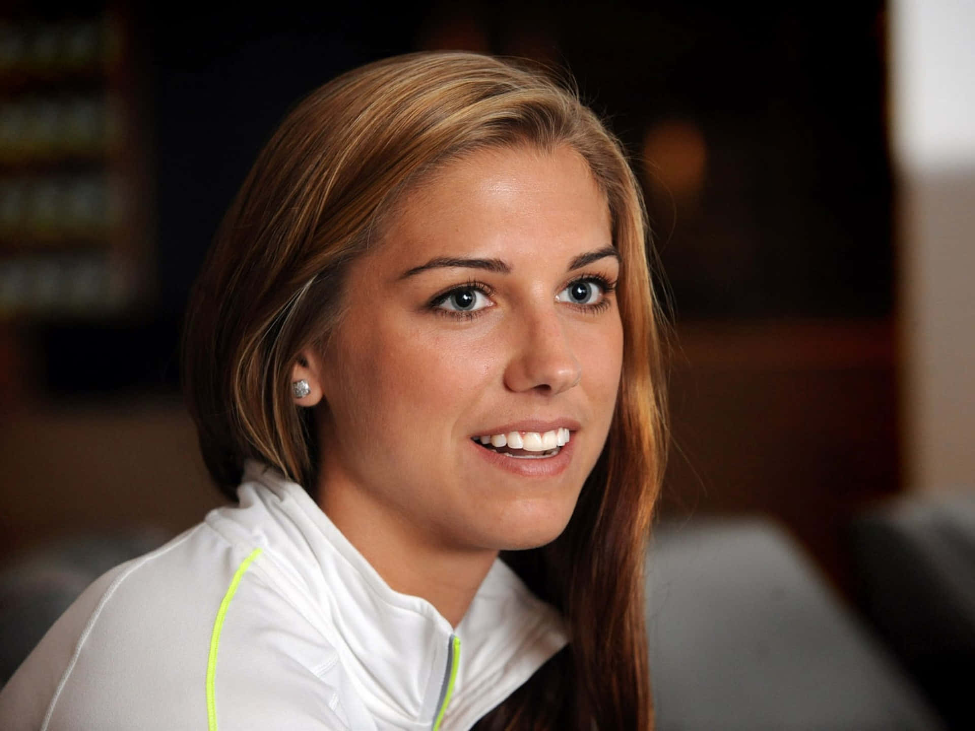 Alex Morgan 桌布 壁紙 — 一位金髮微笑女士的近距離肖像，身穿帶有螢光綠色點綴的白色運動外套。