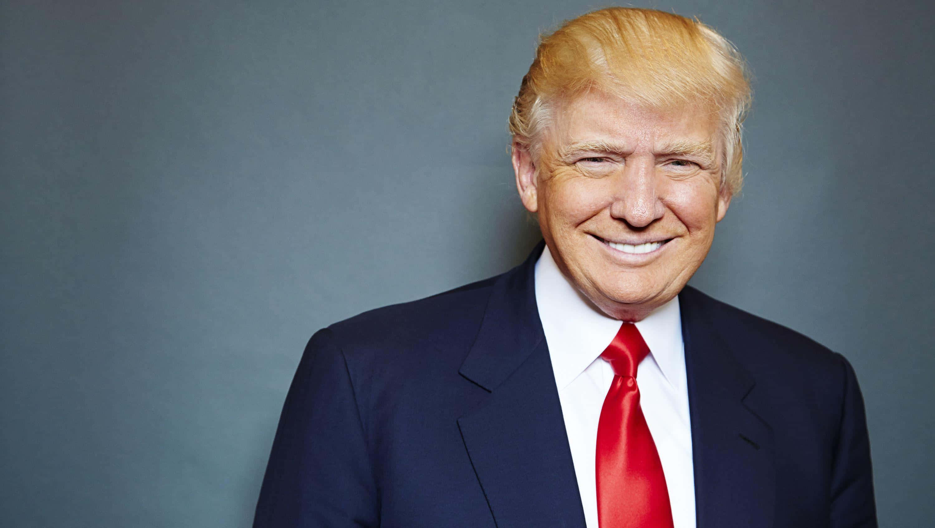 trump 桌布 壁紙 — 一位金髮微笑男士穿著深色西裝和鮮紅色領帶，背景為灰色攝影棚。