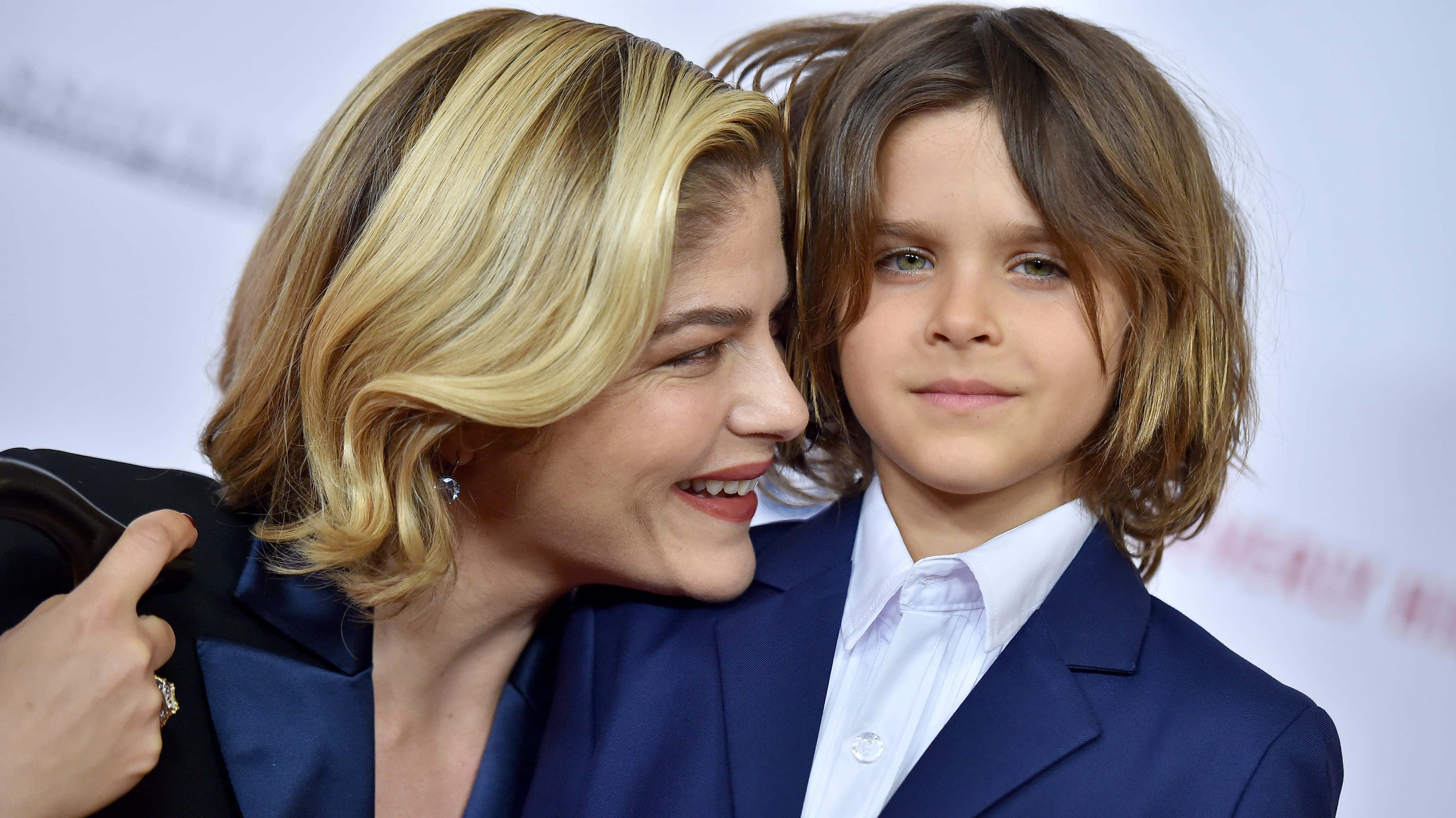 Selma Blair 桌布 壁紙 — 一位金髮微笑的女性擁抱著一個穿著相配藍色西裝的小男孩，背景是模糊的活動場景。