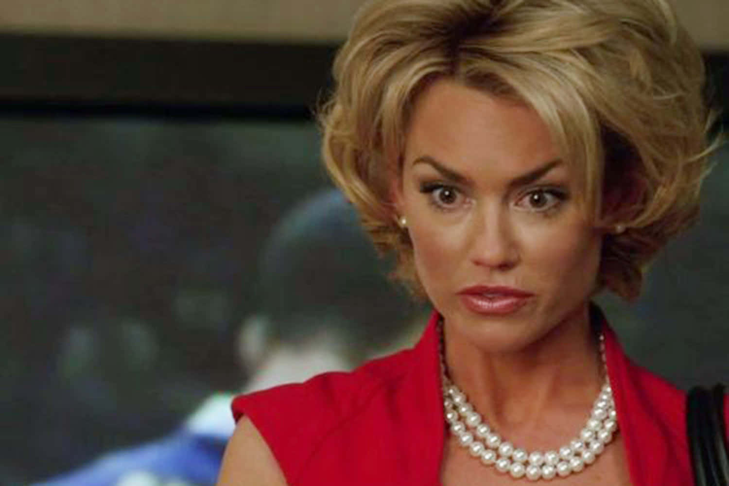 Kelly Carlson 桌布 壁紙 — 一位金髮戴珍珠項鍊的女性，穿著紅色上衣，在模糊背景前專注凝視。