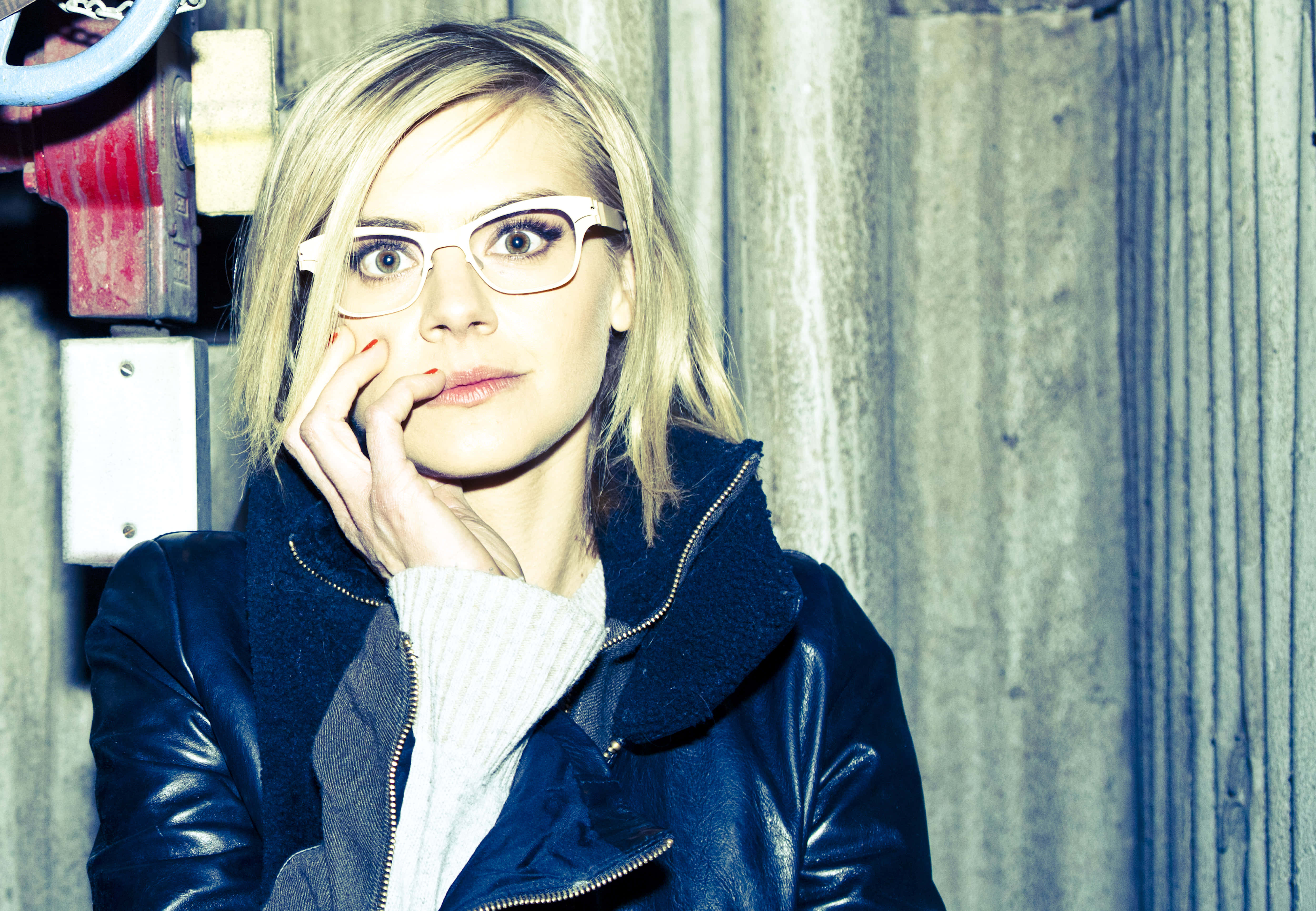 eliza coupe 桌布 壁紙 — 一位金髮戴眼鏡的女性，將下巴擱在手上，靠著風化的木牆，身穿深色皮褸。