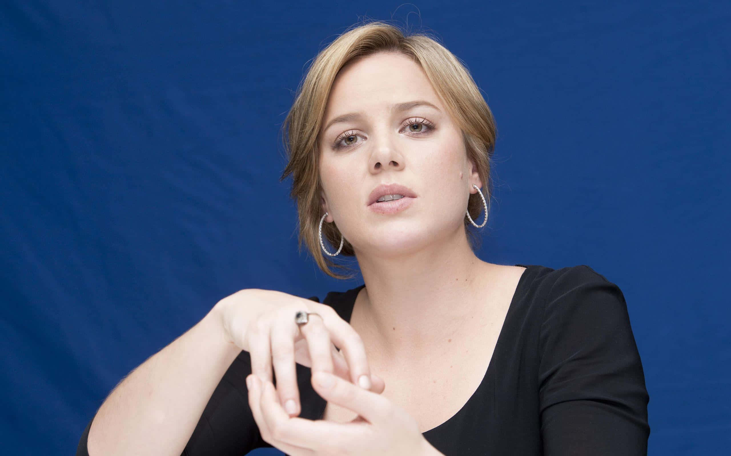 Abbie Cornish 桌布 壁紙 — 一位金髮戴著圈圈耳環的女性，穿著黑色上衣，在純藍色背景下若有所思地凝視著。