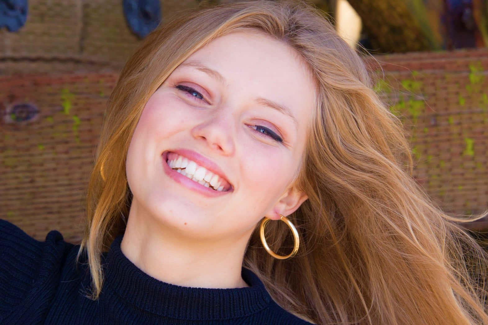 Grace Van Dien 桌布 壁紙 — 一位金髮戴金色圓環耳環的快樂年輕女子，在紋理磚牆背景下燦爛微笑。