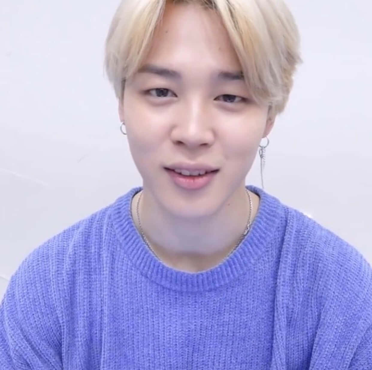 Jimin Aesthetic 桌布 壁紙 — 一位金髮戴銀耳環的年輕男子，穿著質感紫色毛衣，在純白背景前溫柔微笑。