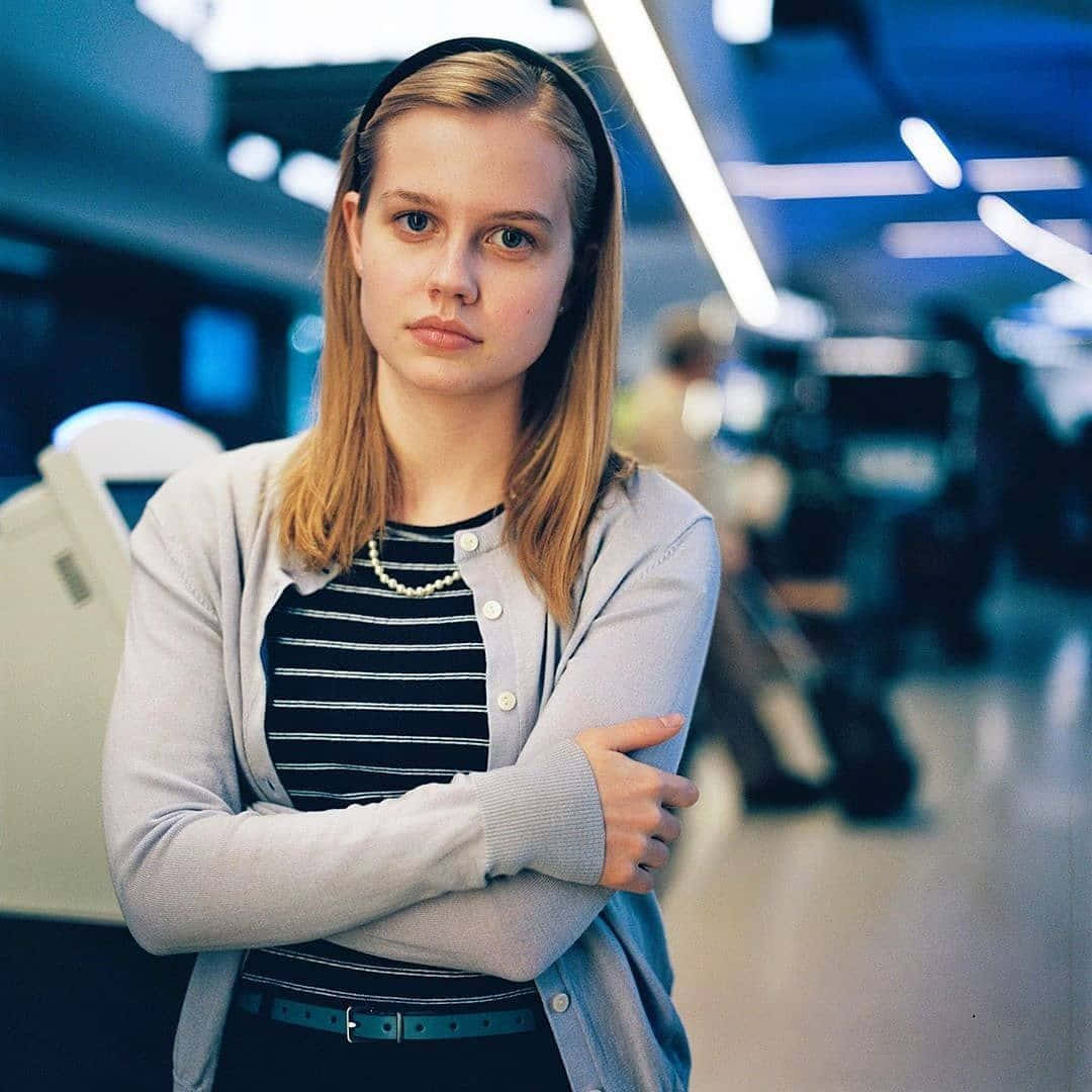 Betty Brant 桌布 壁紙 — 一位金髮戴頭帶的年輕女性，在藍色燈光和模糊同事的現代辦公室中交叉雙臂站立。