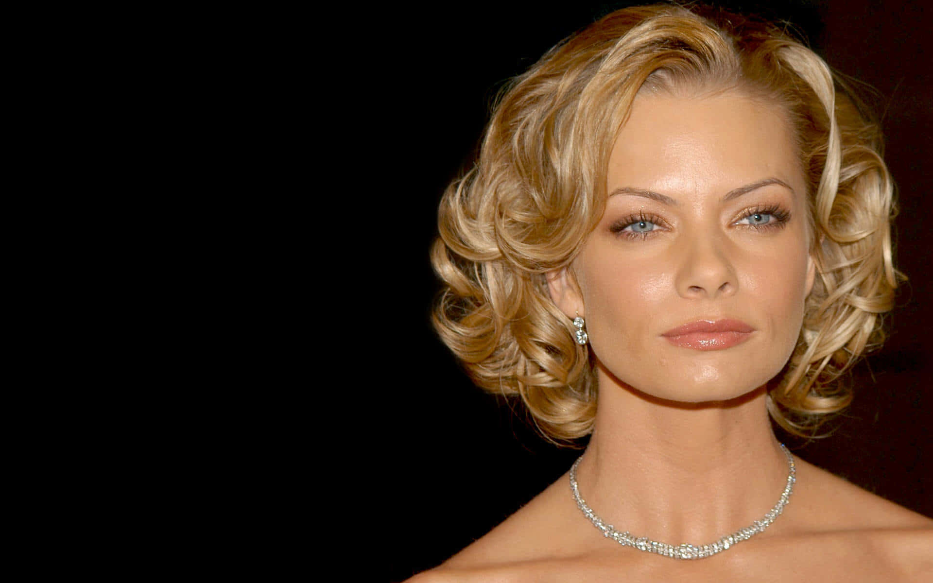 Jaime Pressly 桌布 壁紙 — 一位金髮捲曲、佩戴鑽石珠寶的迷人女性在純黑背景前擺姿勢。