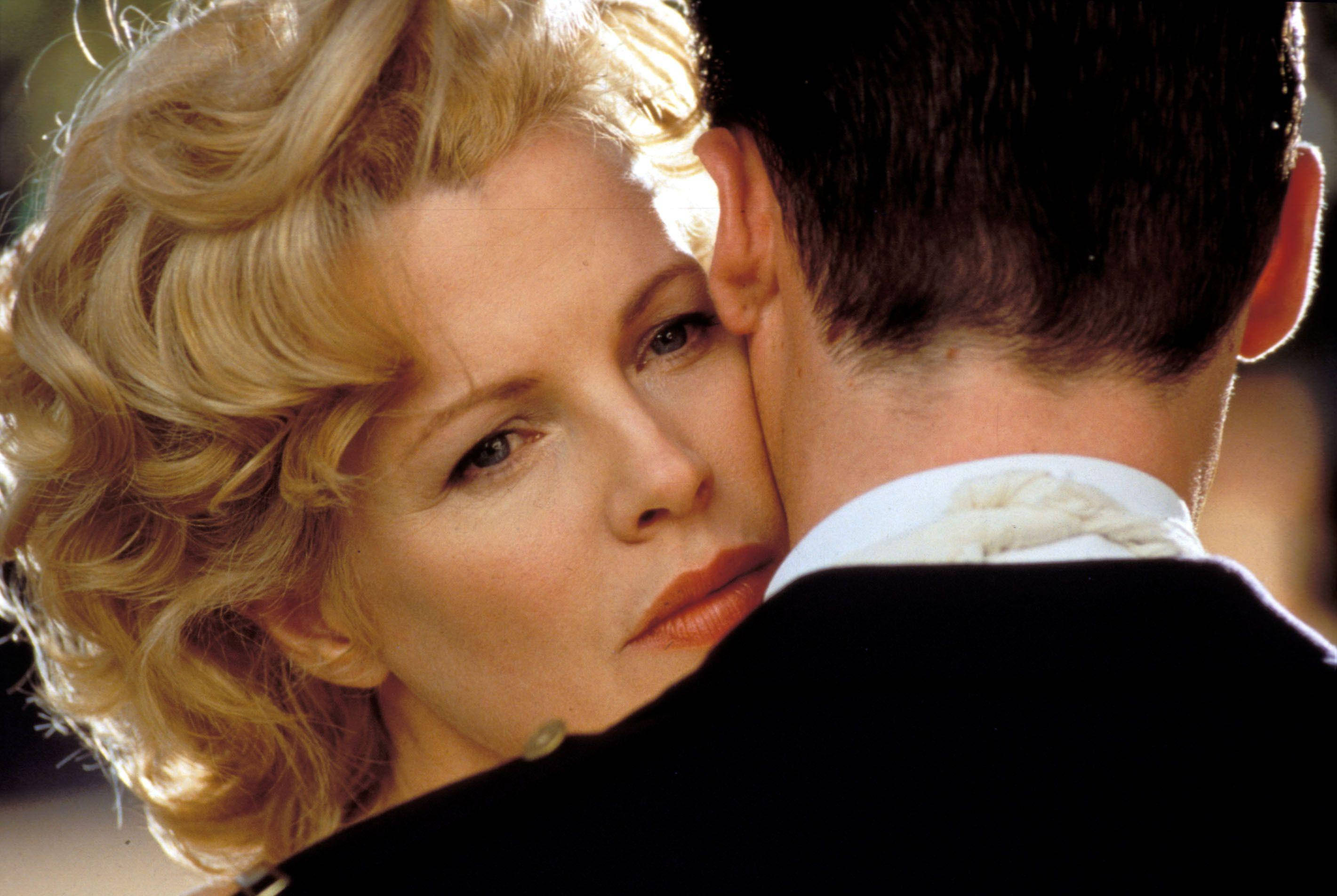 Kim Basinger With Guy Pearce 壁紙 — 一位金髮曲髮女性若有所思地凝視著，同時從後擁抱一位身穿深色西裝的男士。