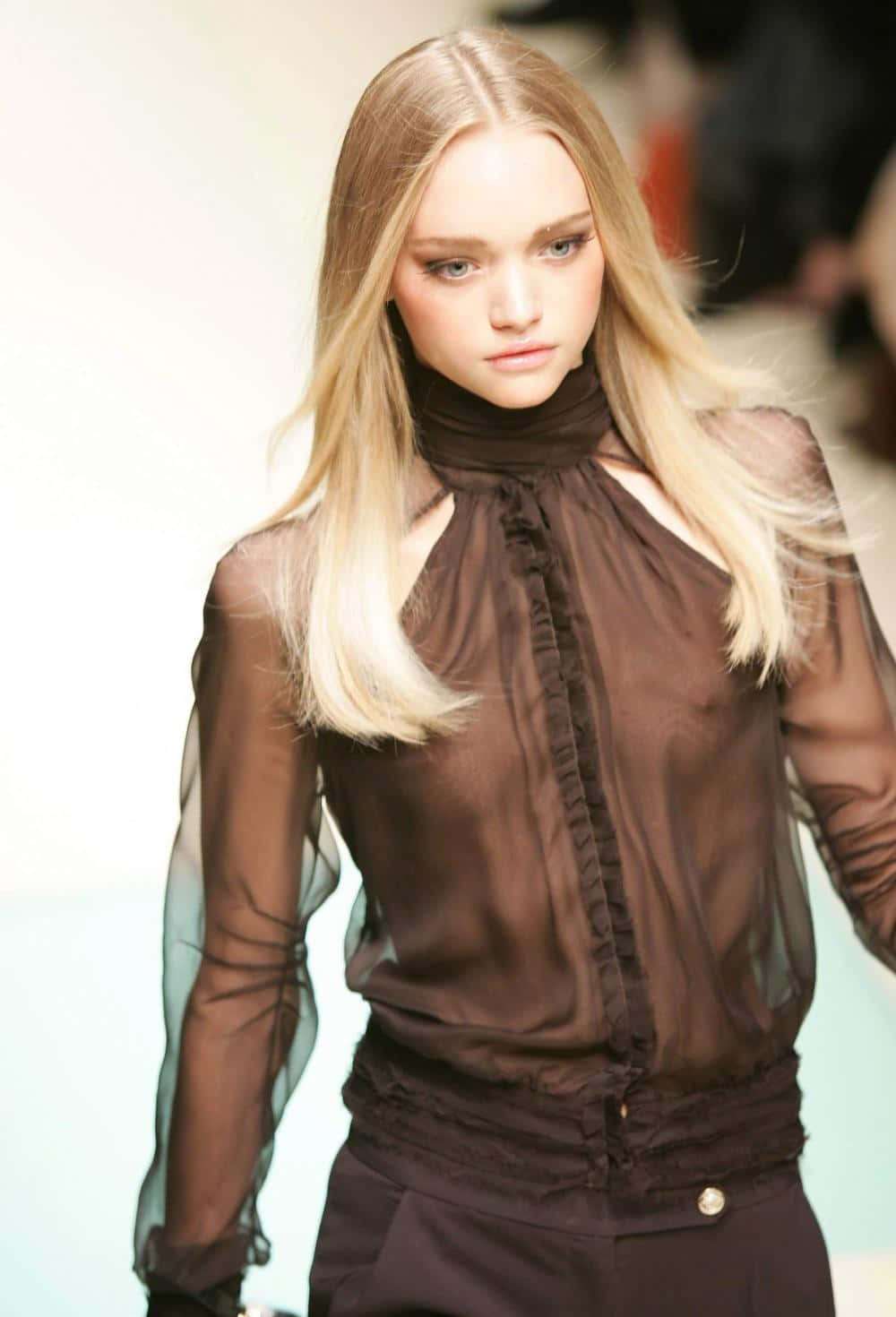 Gemma Ward 桌布 壁紙 — 一位金髮模特兒自信地走在天橋上，身穿帶荷葉邊細節的透視棕色襯衫和配套褲子。