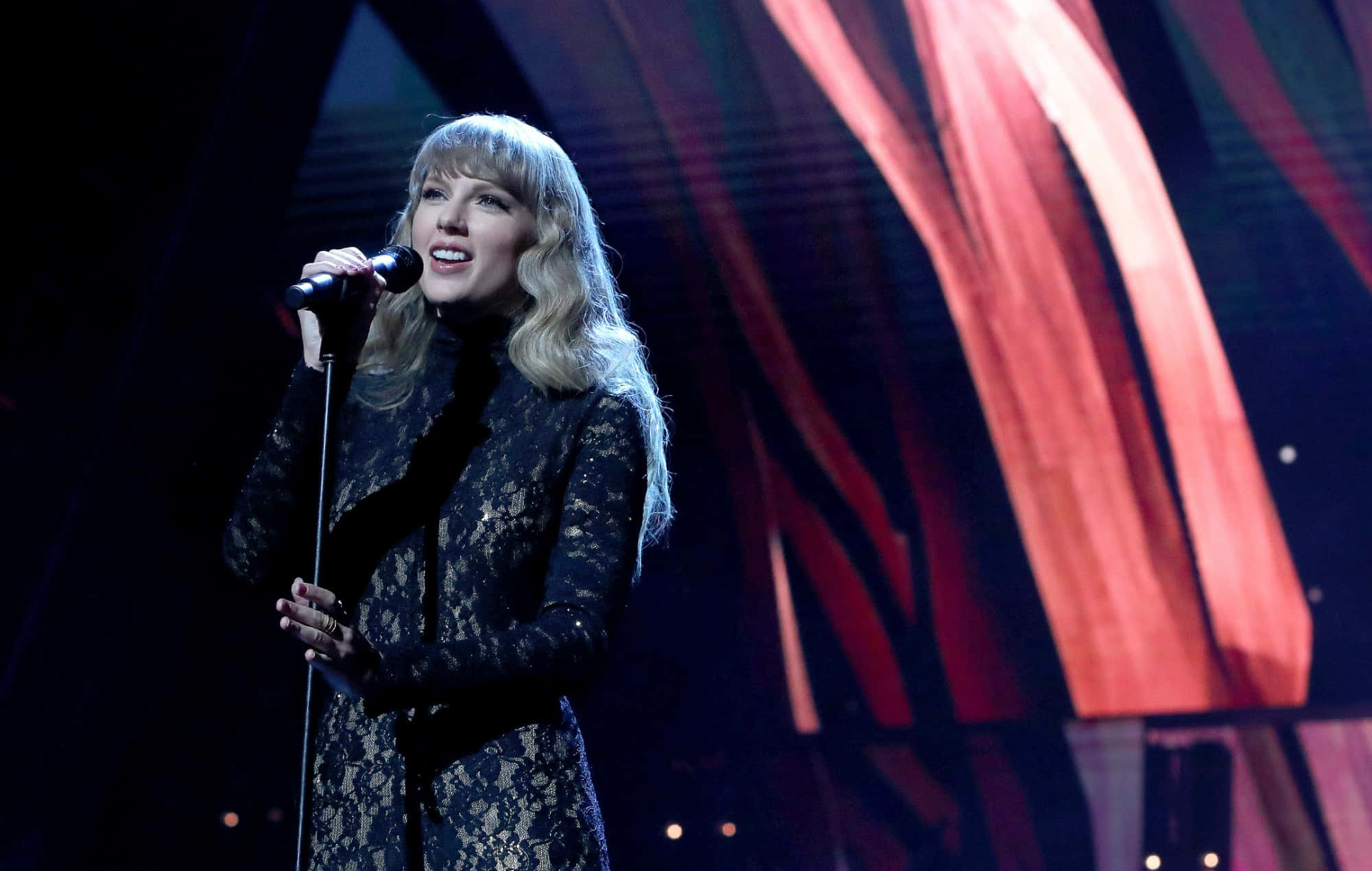 Taylor Swift 桌布 壁紙 — 一位金髮歌手穿著黑色蕾絲服裝，在戲劇性的紅色舞台燈光下熱情地對著麥克風表演。