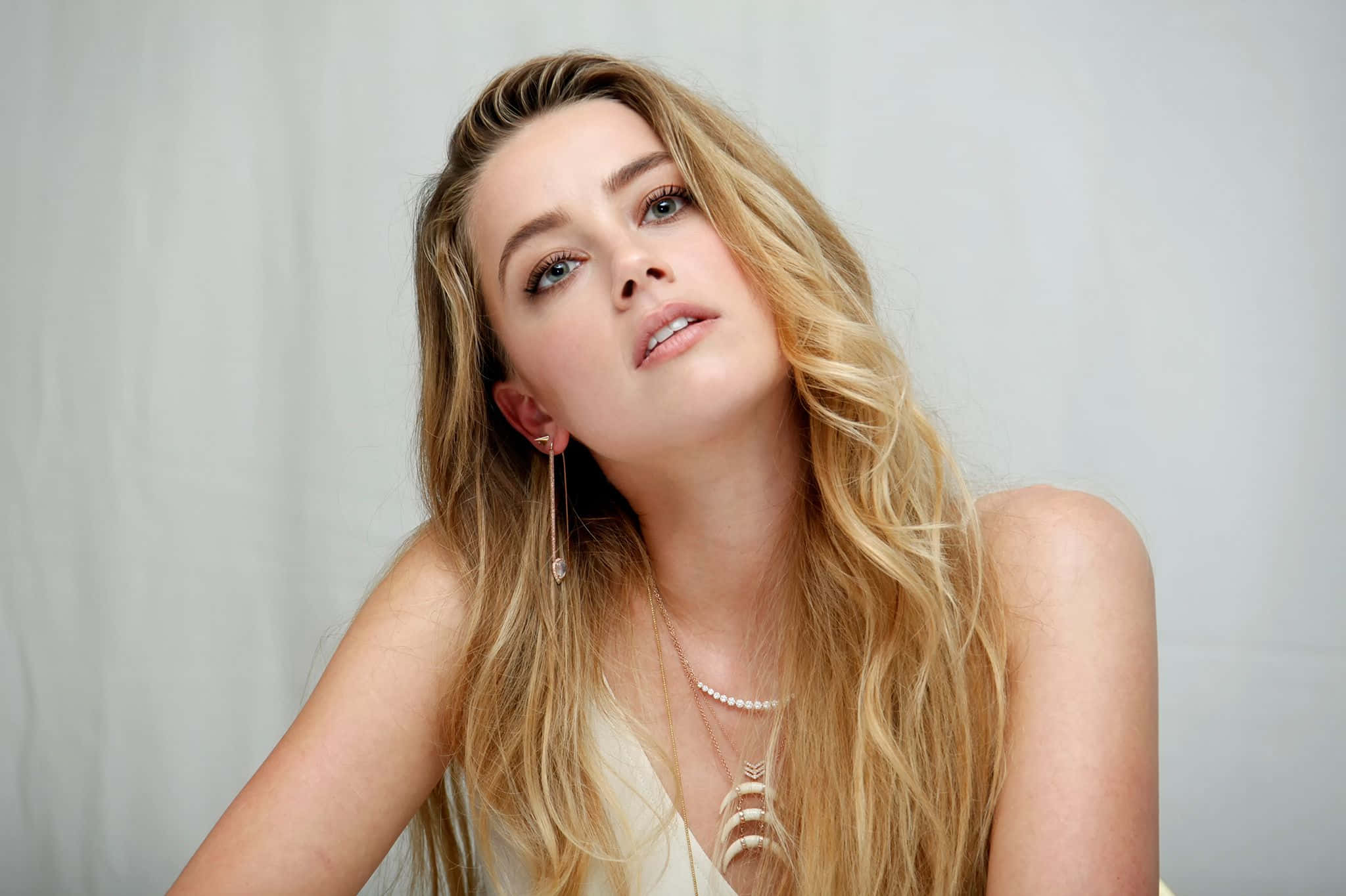 Amber Heard 桌布 壁紙 — 一位金髮波浪長髮的女性，佩戴精緻珠寶，在純淺灰色背景下向上凝視。