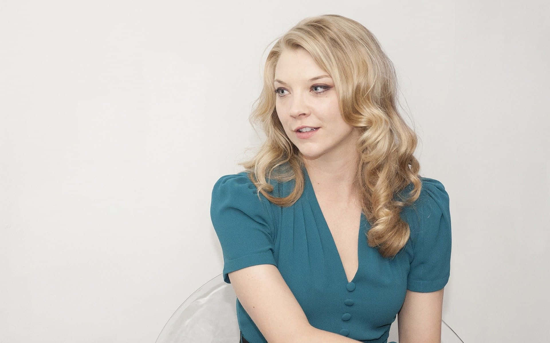 natalie dormer 桌布 壁紙 — 一位金髮波浪長髮的女性，身穿青綠色連衣裙，坐在透明椅子上，背景是純白色。