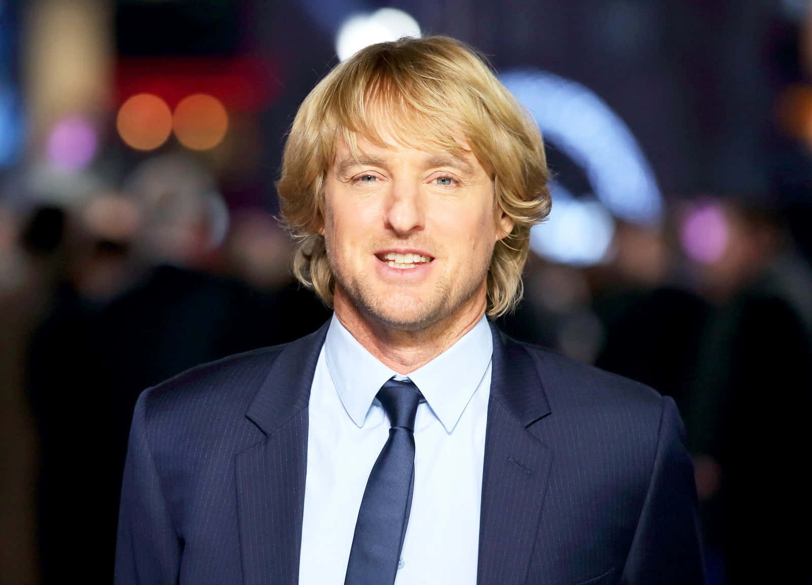 Owen Wilson 桌布 壁紙 — 一位金髮男子身穿深色西裝，在模糊的城市夜景背景下，對著鏡頭微笑，背景有閃爍的燈光。