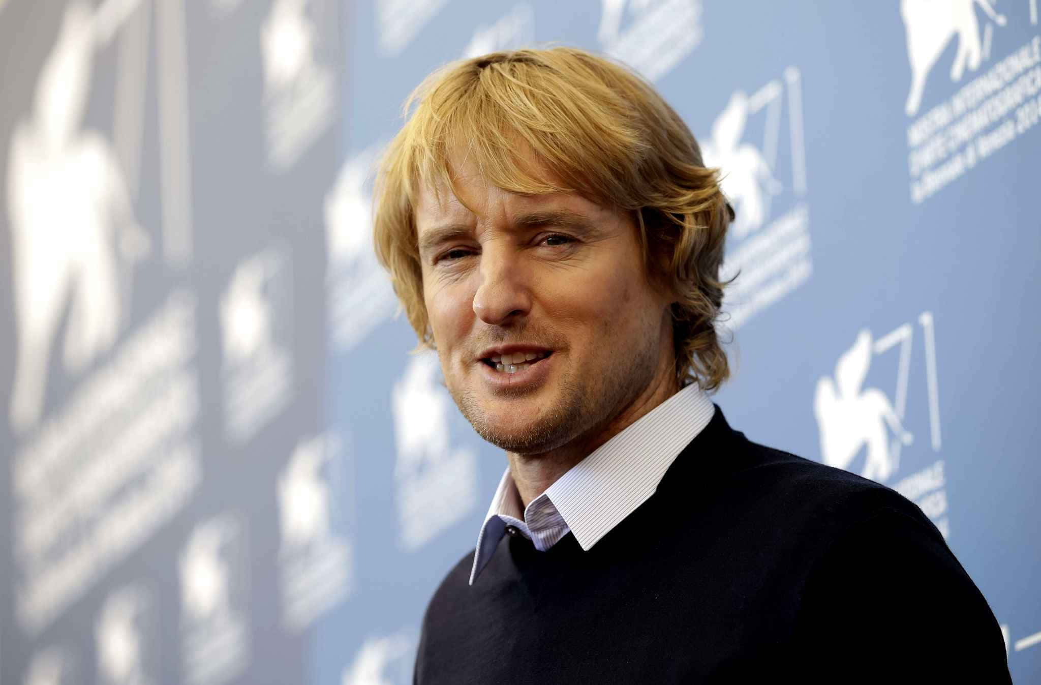 Owen Wilson 桌布 壁紙 — 一位金髮男子面帶微笑，站在帶有白色標誌的藍色背景前。