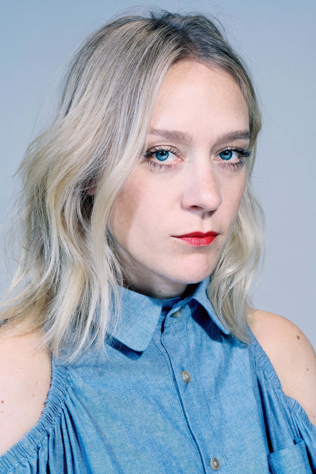 Chloë Sevigny 桌布 壁紙 — 一位金髮碧眼、擁有迷人藍色眼睛的女性，身穿藍色露肩襯衫，塗著大膽的紅色唇膏，在柔和的灰色背景前直視鏡頭。