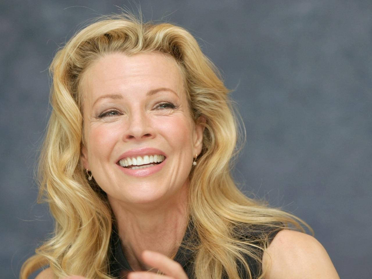 Actress Kim Basinger At Beverly Hills 2006 壁紙 — 一位金髮碧眼、笑容燦爛、波浪長髮的女性，在柔和的灰色背景前。