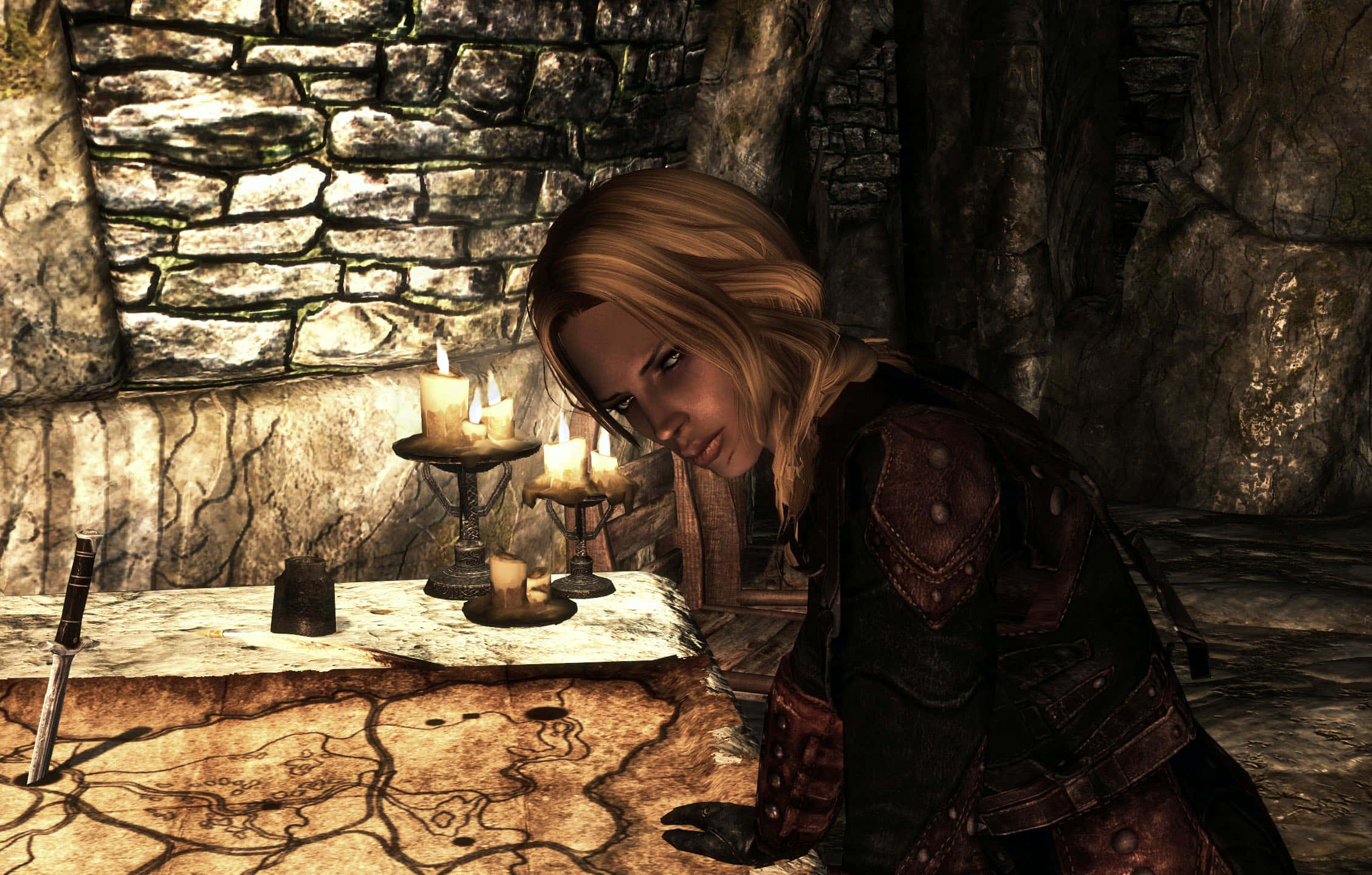 Skyrim Astrid 桌布 壁紙 — 一位金髮穿甲的女性在昏暗的地牢中，俯身在一張石桌上，桌上放著發光的蠟燭和地圖。