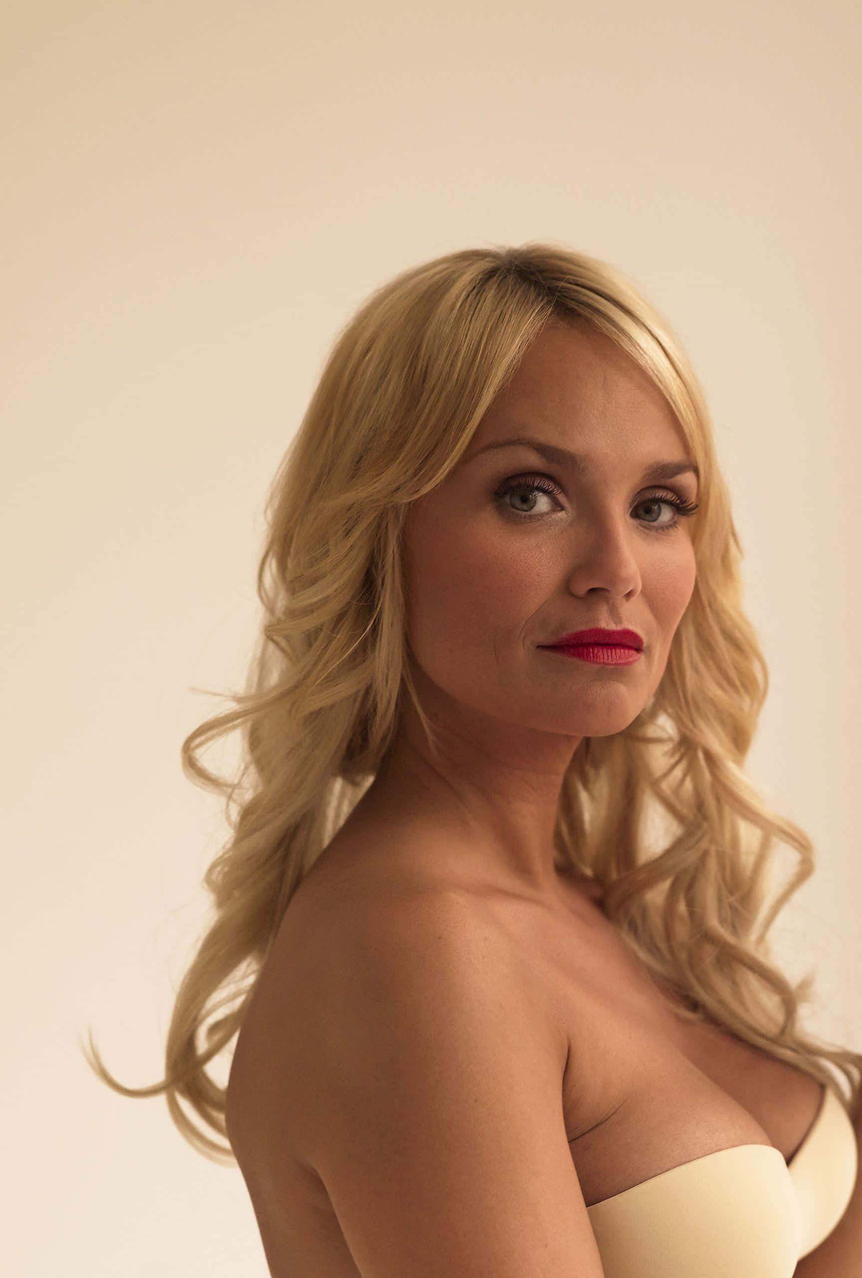 kristin chenoweth 桌布 壁紙 — 一位金髮紅唇、波浪長髮的女性，身穿米白色無肩帶連衣裙，在柔和的米色背景前擺姿勢。