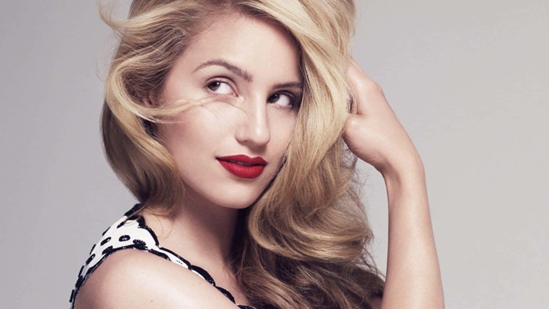 dianna agron 桌布 壁紙 — 一位金髮紅唇、波浪長髮的女性在純灰色背景前擺姿勢，手輕輕觸碰頭髮。