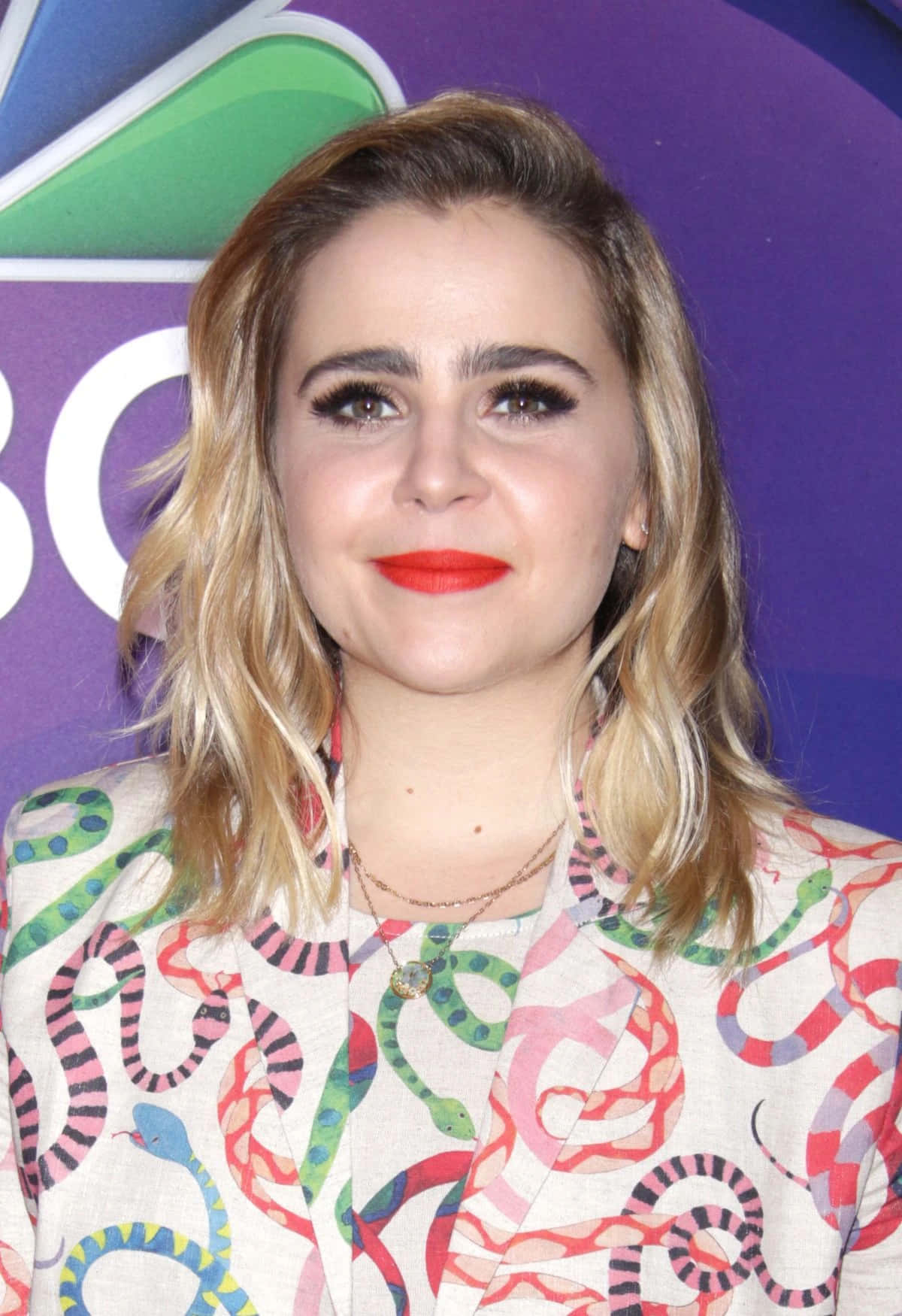 Mae Whitman 桌布 壁紙 — 一位金髮紅唇的女性，穿著色彩繽紛的蛇紋外套，在紫色背景前微笑。