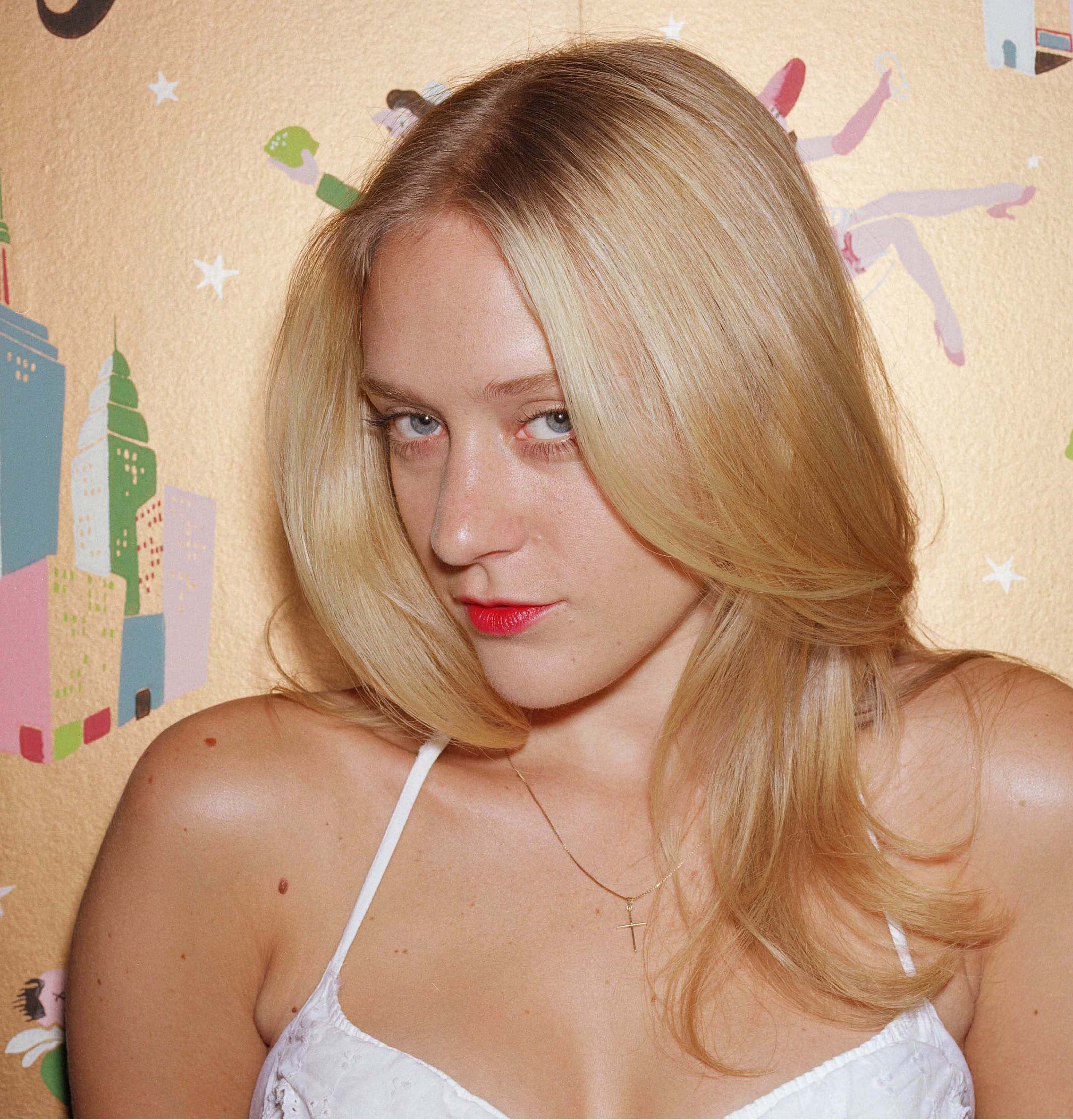 Chloë Sevigny 桌布 壁紙 — 一位金髮紅唇的女性在夢幻插畫壁紙背景下直視鏡頭。