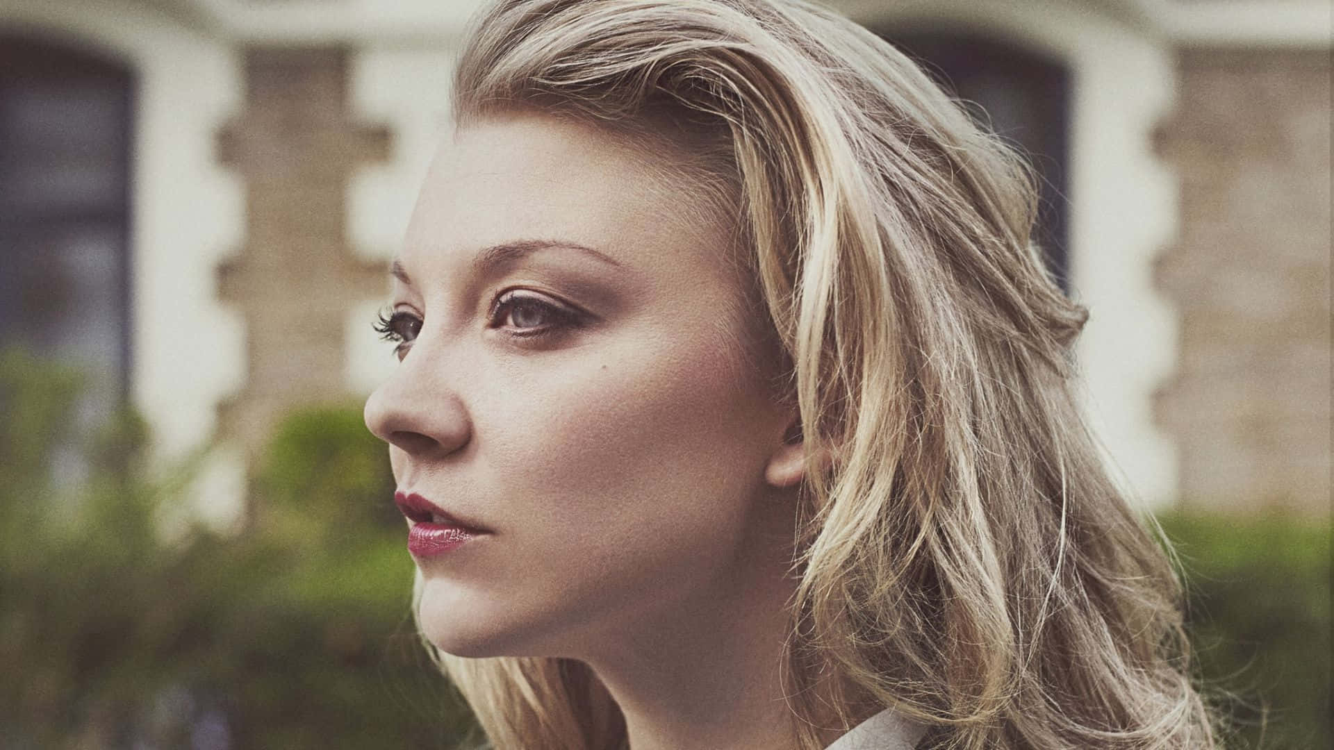 natalie dormer 桌布 壁紙 — 一位金髮紅唇的女性在戶外，以模糊的磚建築為背景，若有所思地凝視著。