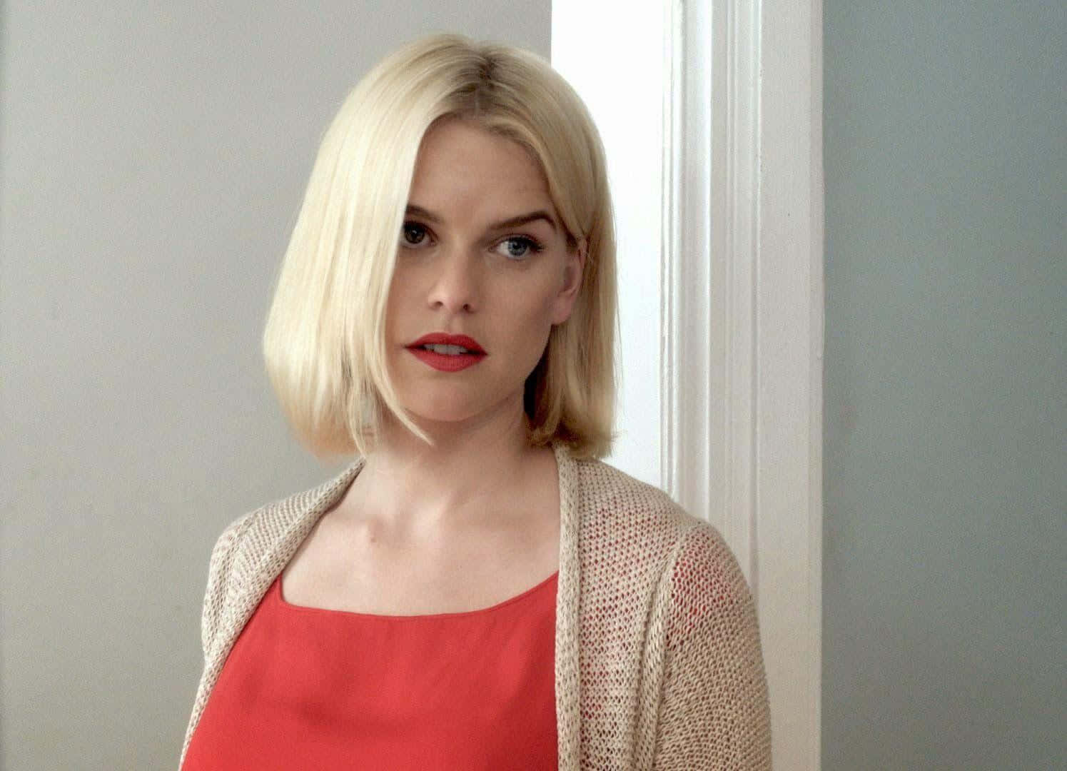 alice eve 桌布 壁紙 — 一位金髮紅唇的女性穿著紅色上衣和米色開襟毛衣，站在白色門框旁。
