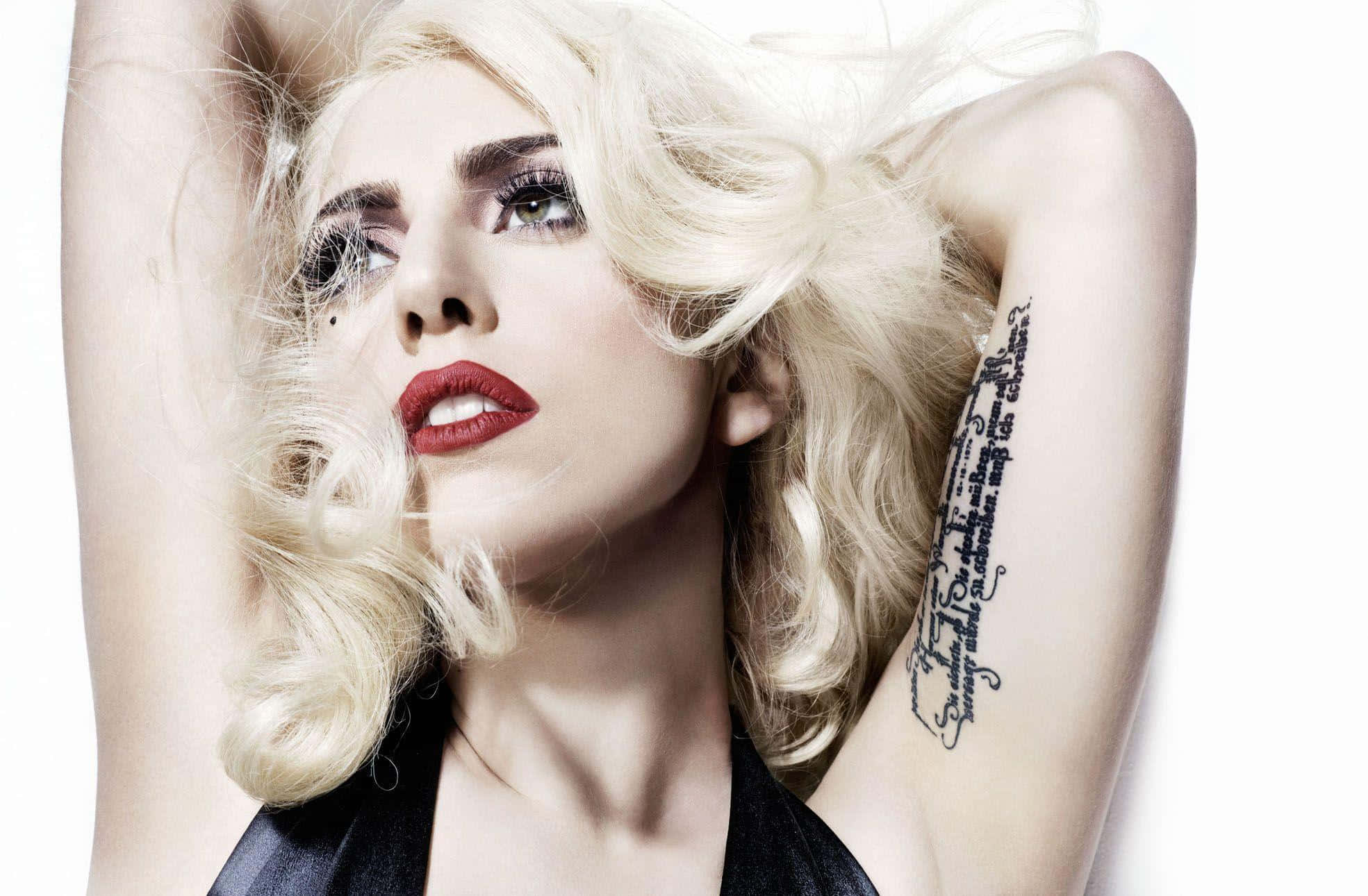 Lady Gaga 桌布 壁紙 — 一位金髮美女，有著大膽的紅唇和手臂上的黑色紋身，在純白背景前擺姿勢。