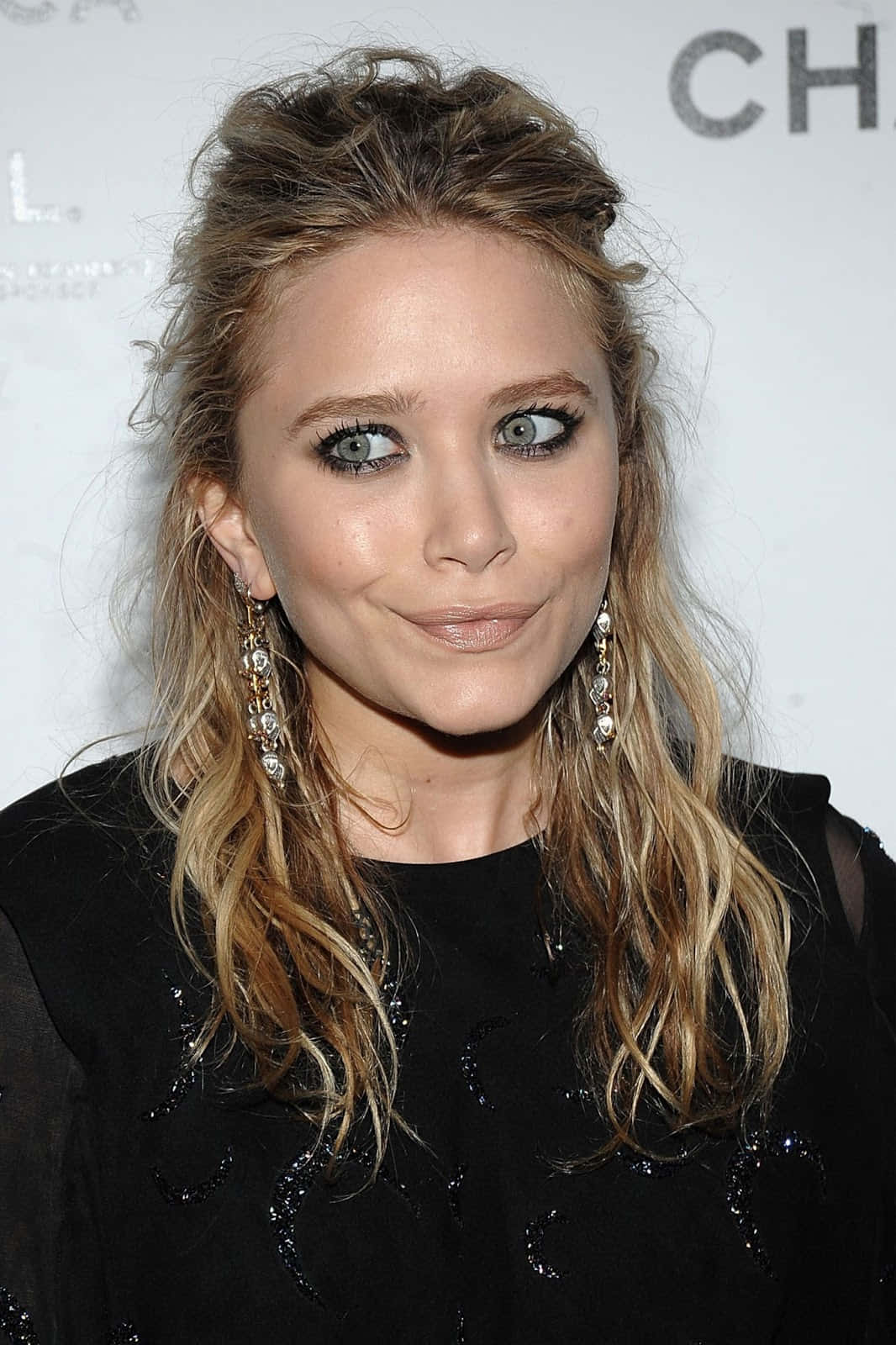 Mary Kate Olsen 桌布 壁紙 — 一位金髮蓬鬆、擁有引人注目綠眼睛的女子的特寫肖像，佩戴優雅的垂墜耳環和黑色裝飾上衣。
