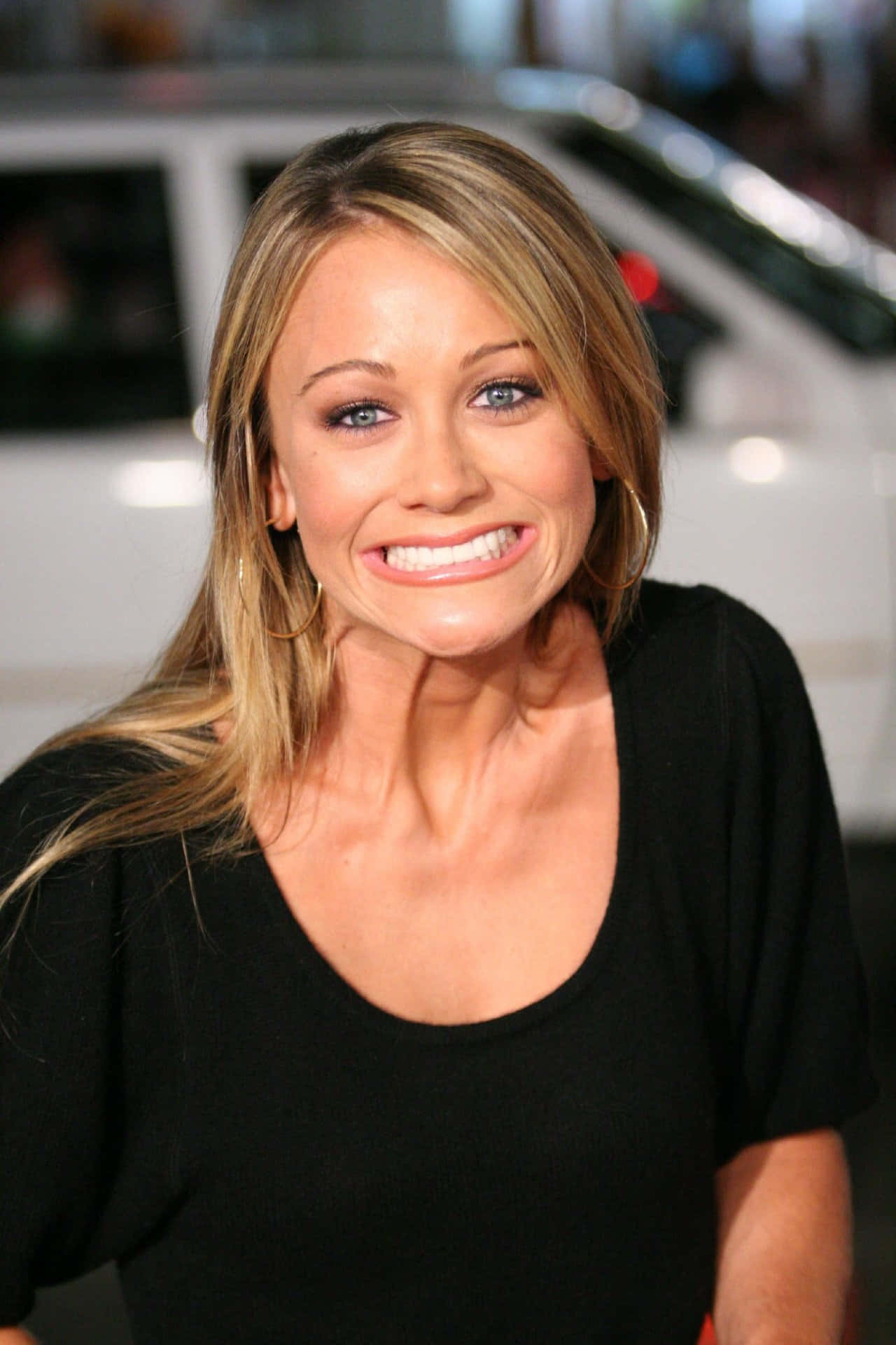 Christine Taylor 桌布 壁紙 — 一位金髮藍眼、面帶微笑的女性，穿著黑色上衣，背景是模糊的汽車。