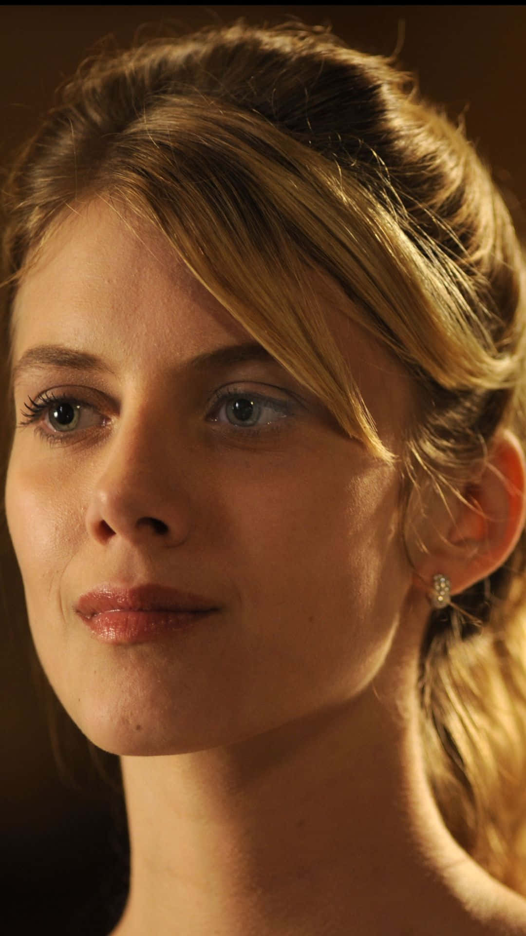 Mélanie Laurent 桌布 壁紙 — 一位金髮藍眼女士在溫暖金色燈光下沉思凝視的特寫肖像。