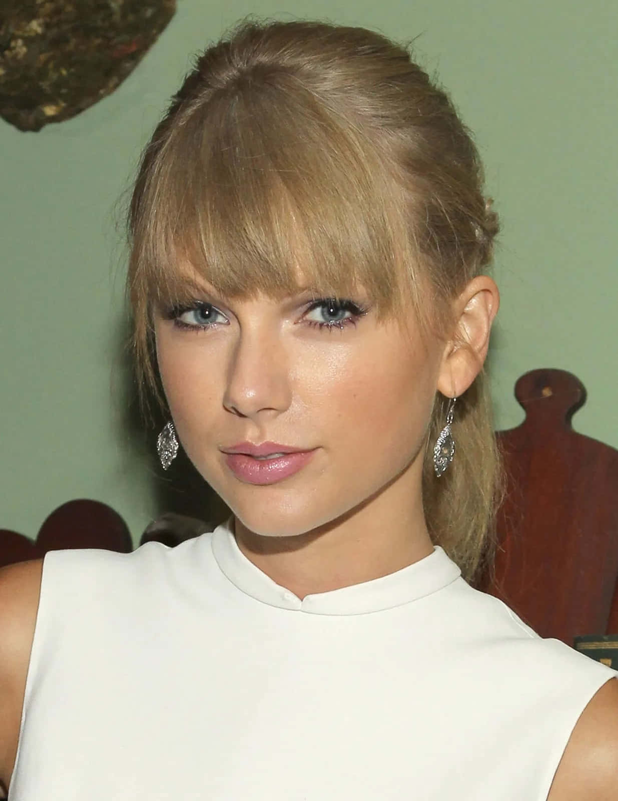 Taylor Swift 桌布 壁紙 — 一位金髮藍眼女士身穿白色無袖上衣和垂墜耳環的特寫肖像。