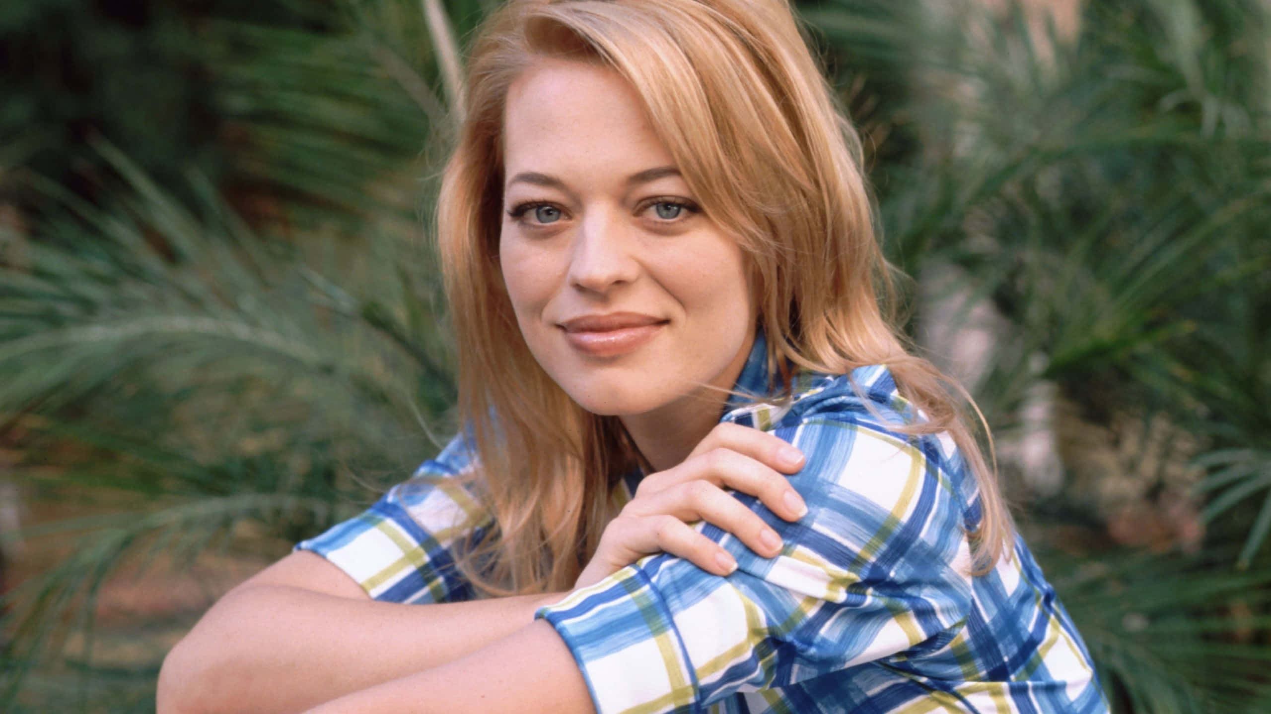 Jeri Ryan 桌布 壁紙 — 一位金髮藍眼的女性，穿著藍色格仔襯衫，在模糊的綠色棕櫚樹背景下溫柔微笑。