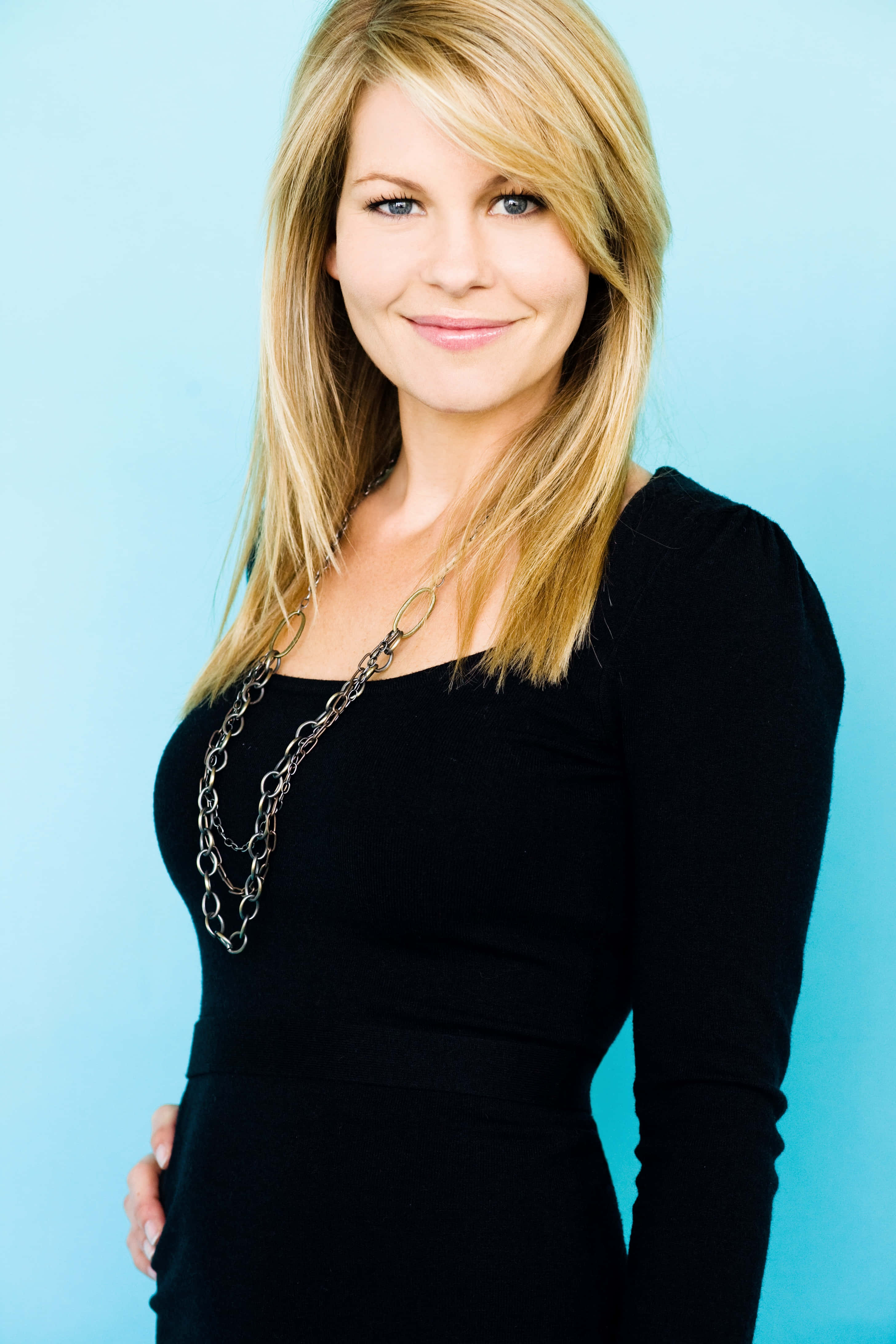 candace cameron bure 桌布 壁紙 — 一位金髮藍眼的女性，穿著黑色連衣裙和銀色鏈條項鍊，在純藍色背景前溫柔微笑。