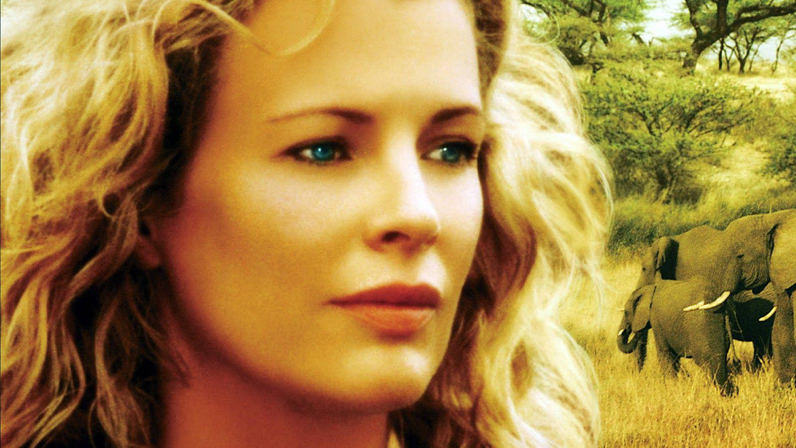 Striking Kim Basinger as Kuki Gallmann 壁紙 — 一位金髮藍眼的女性向前凝視，身後大象在陽光照耀的草原上行走。
