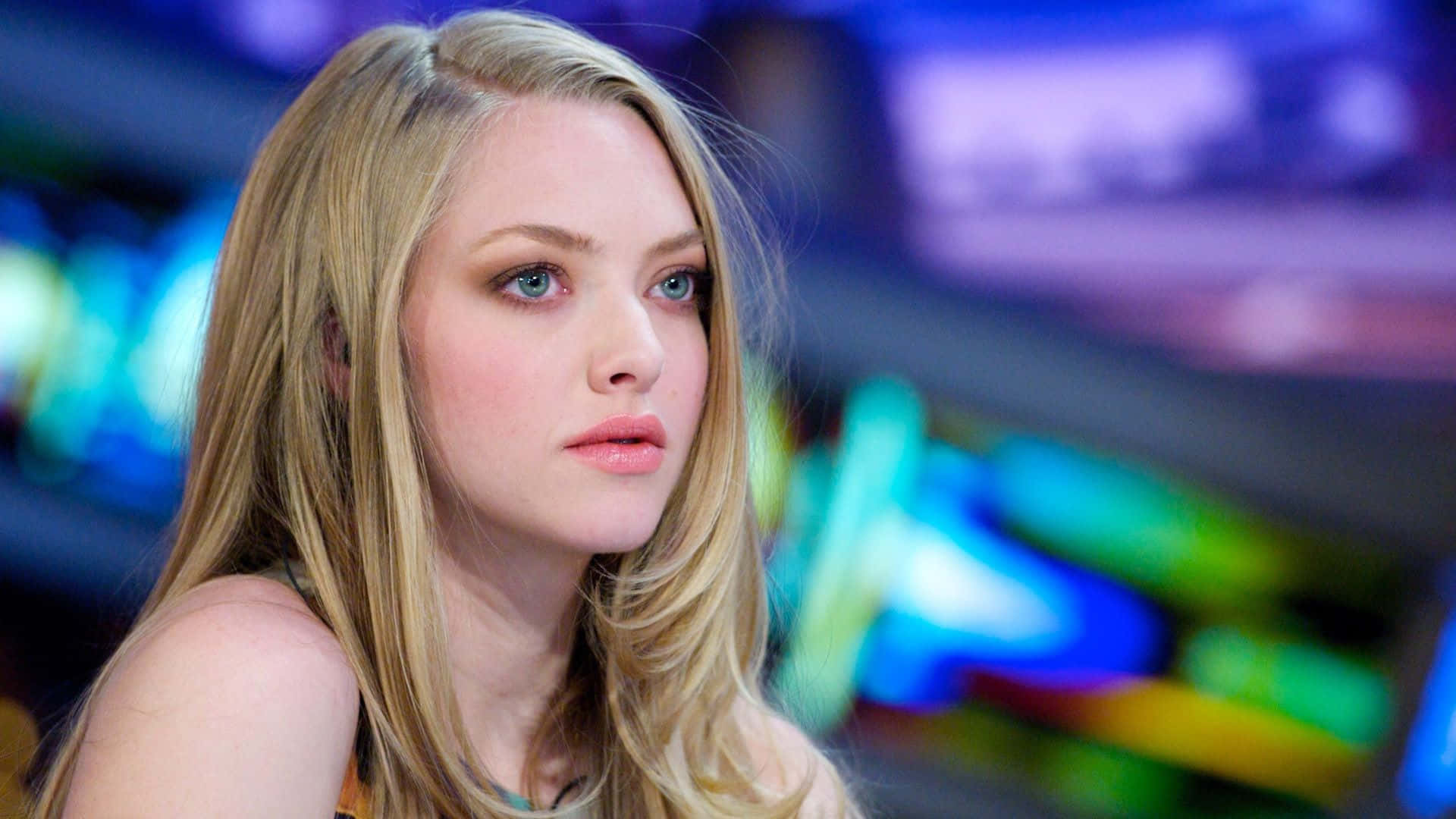 Amanda Seyfried 桌布 壁紙 — 一位金髮藍眼的年輕女子，在充滿活力的模糊霓虹燈背景前若有所思地凝視著。