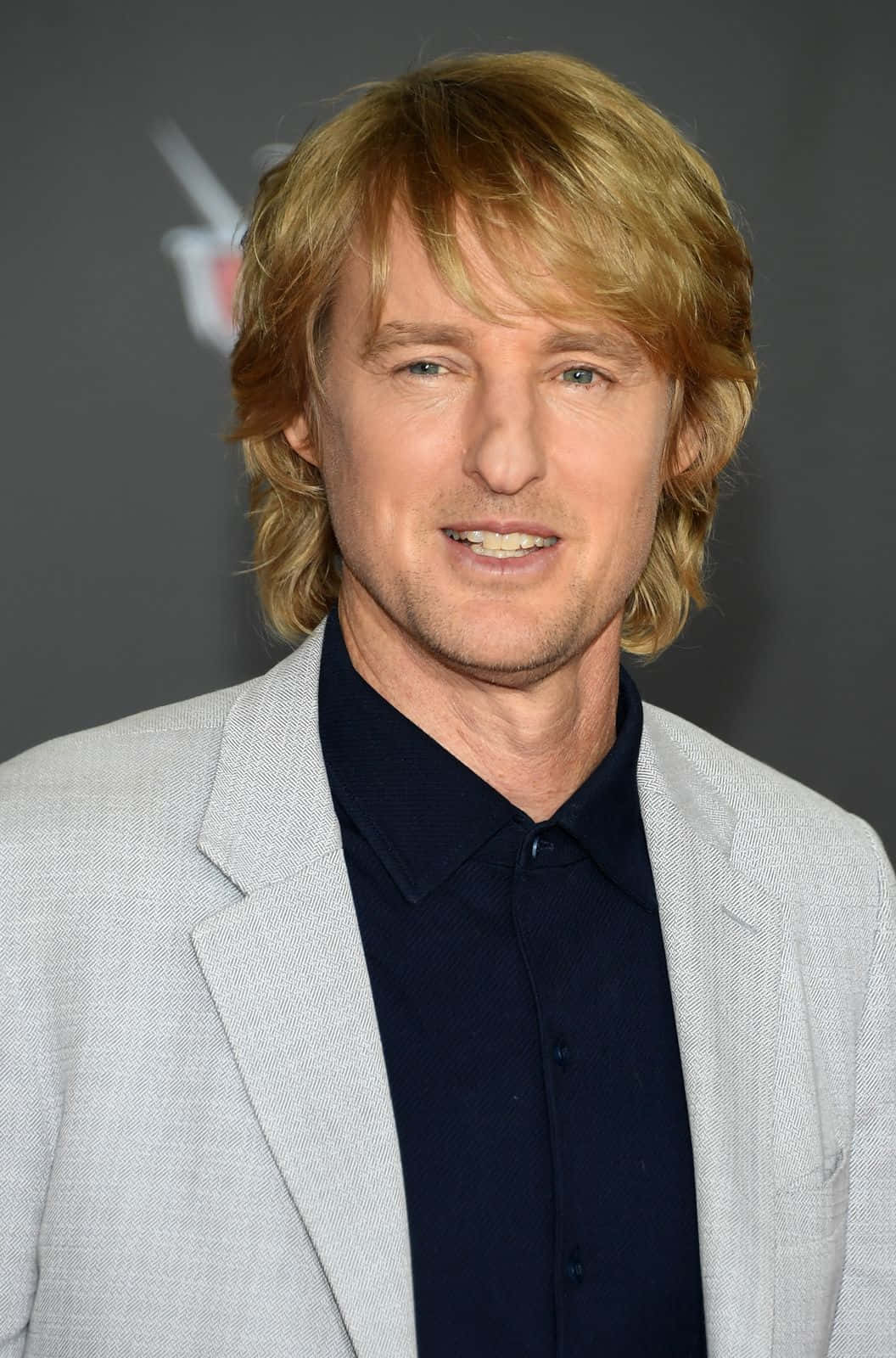 Owen Wilson 桌布 壁紙 — 一位金髮藍眼的男士微笑著，身穿灰色西裝外套，內搭深藍色襯衫。