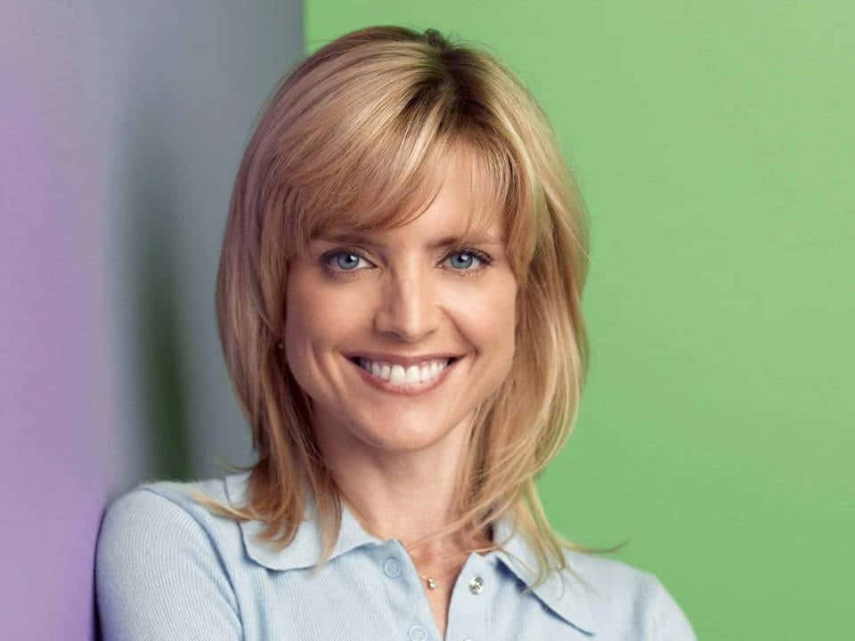 Courtney Thorne Smith 桌布 壁紙 — 一位金髮藍眼的開朗女性，在充滿活力的綠色和紫色背景前溫暖地微笑。