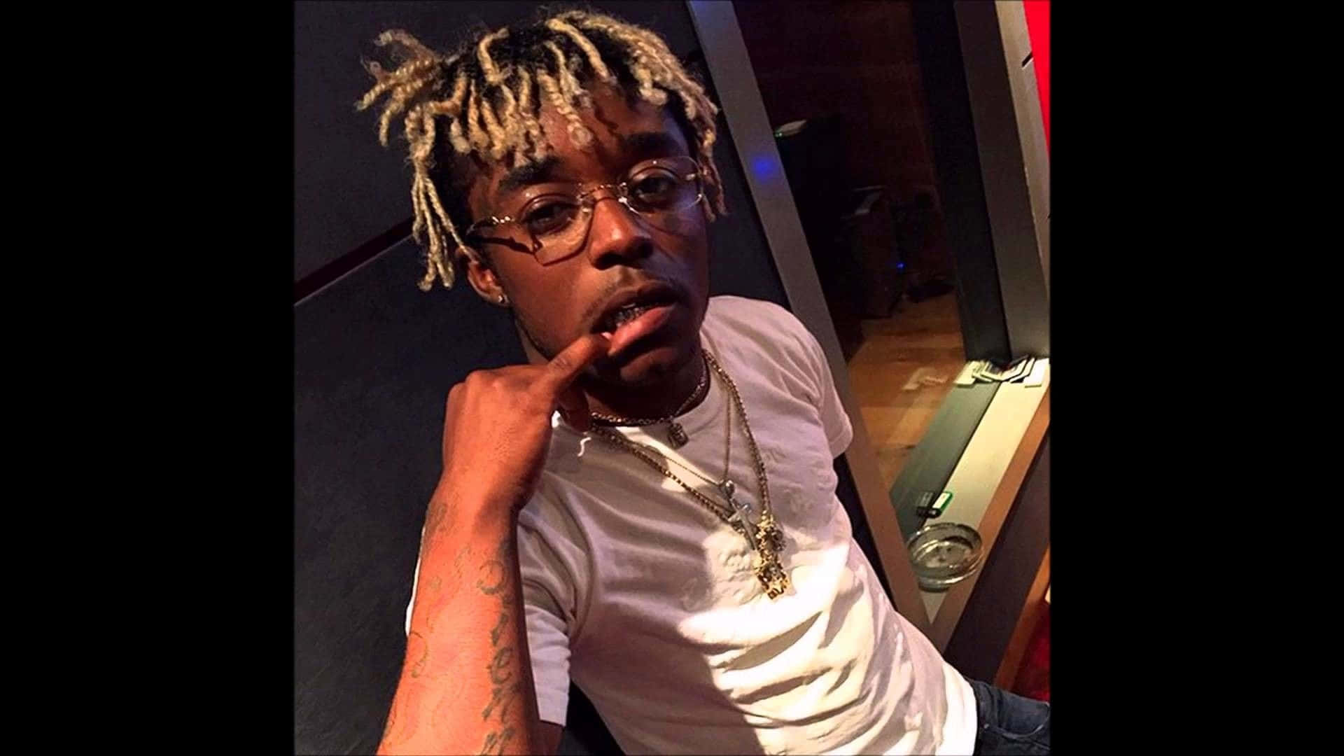 Lil Uzi Vert 桌布 壁紙 — 一位金髮辮子、戴眼鏡的年輕男子在昏暗的房間裡若有所思地擺姿勢，身穿白色T恤和層疊項鍊。