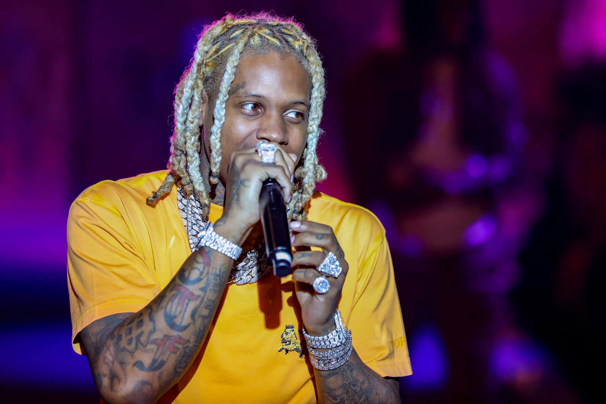 lil durk 桌布 壁紙 — 一位金髮辮子、有紋身的說唱歌手在紫色燈光下穿著黃色襯衫在舞台上表演。