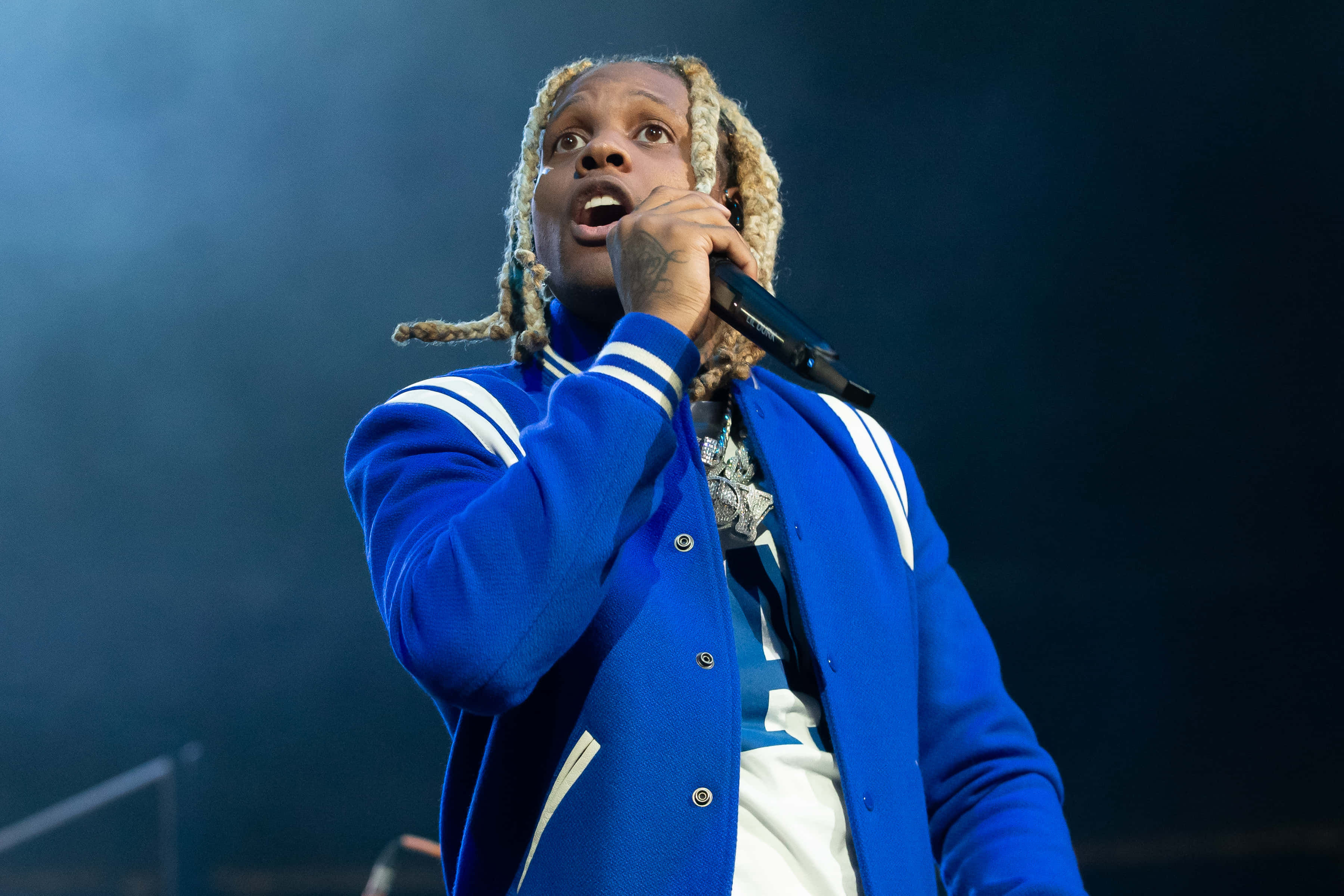 lil durk 桌布 壁紙 — 一位金髮辮子的男饒舌歌手在舞台燈光下穿著藍色大學外套，對著麥克風熱情演唱。