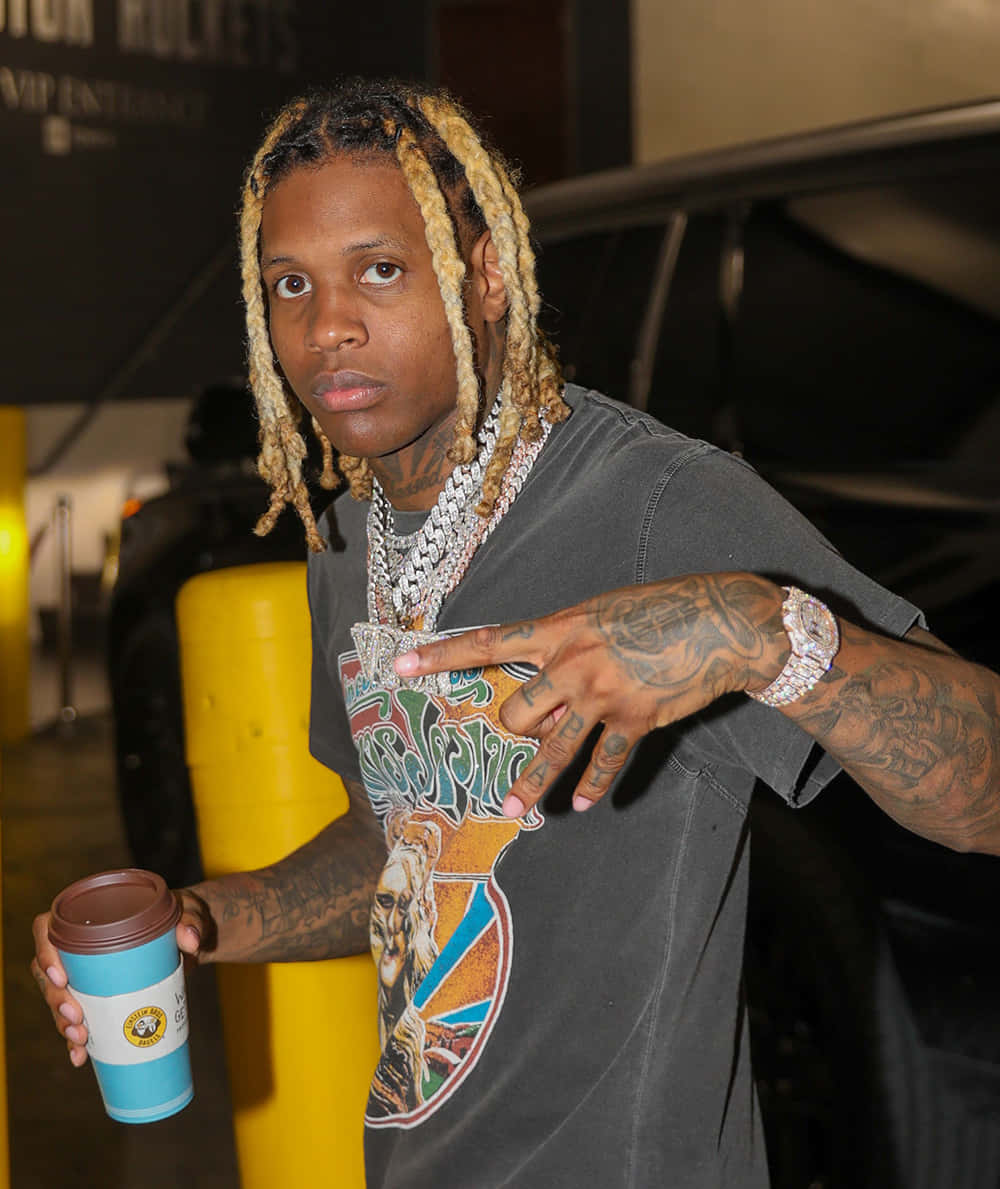 lil durk 桌布 壁紙 — 一位金髮辮子紋身男子在昏暗的室內空間自信地拿著咖啡杯擺姿勢。