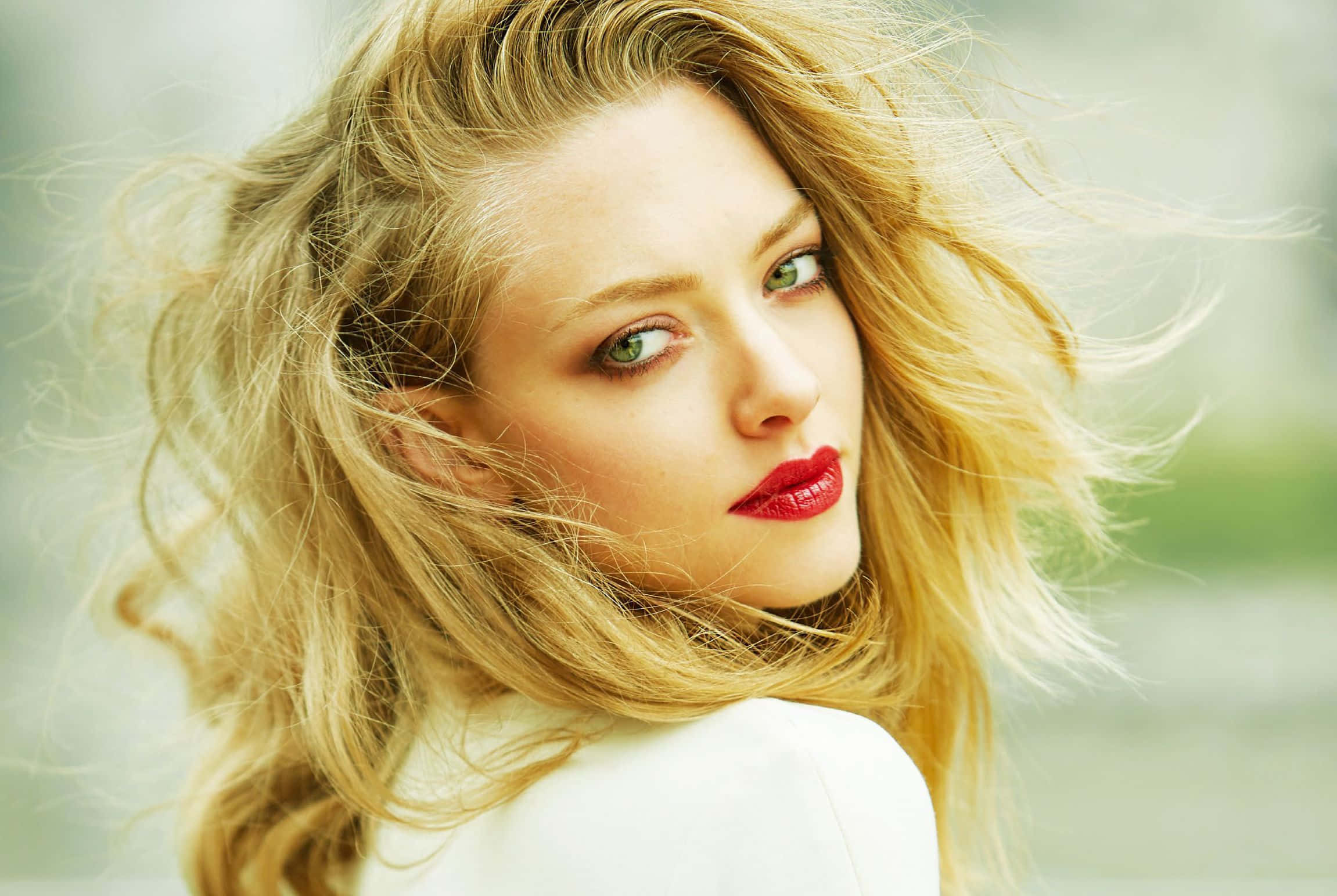 Amanda Seyfried 桌布 壁紙 — 一位金髮隨風飄揚、紅唇大膽的女性，在柔和模糊的背景前凝視著。
