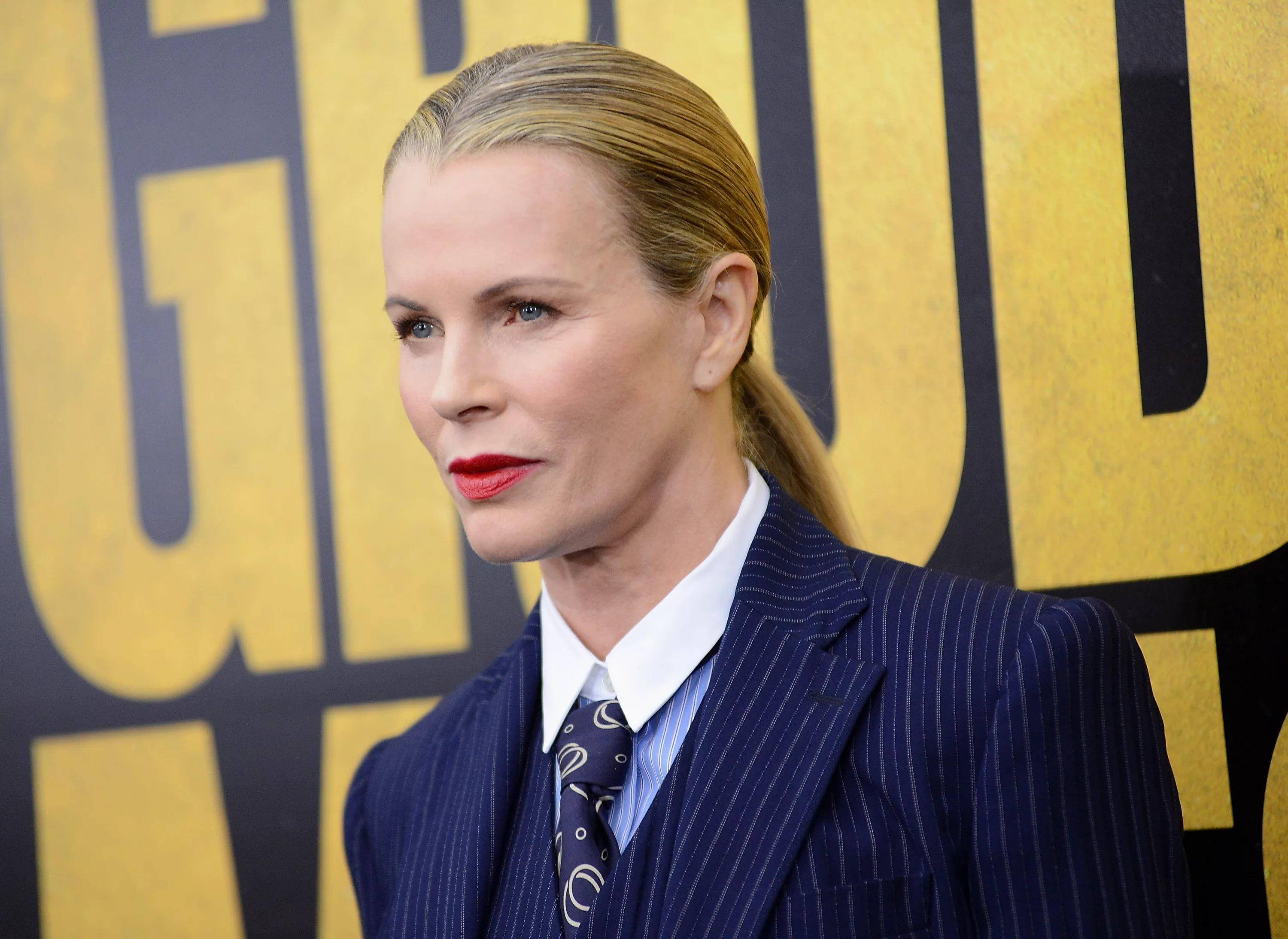 Kim Basinger Grudge Match Premiere 2013 壁紙 — 一位金髮馬尾辮的女性，穿著海軍藍細條紋西裝和紅色口紅，背景為黃色和黑色。